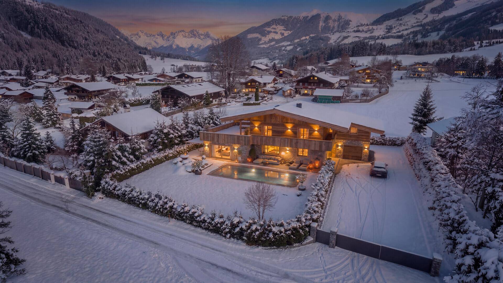 Abendliche Luftaufnahme des beleuchteten Chalets mit Pool und verschneitem Garten in den Bergen.