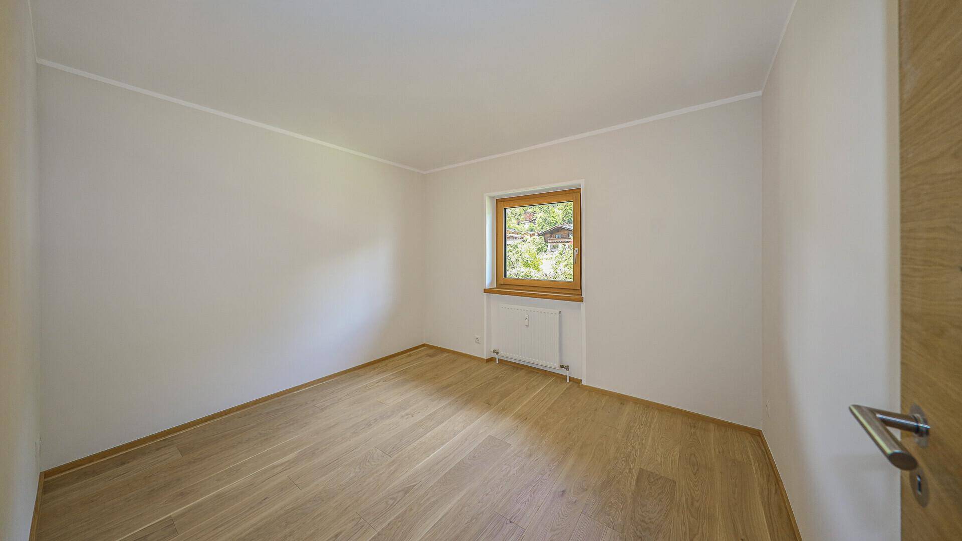 Leeres Schlafzimmer mit Holzboden, weiß gestrichenen Wänden und einem Fenster mit Blick ins Grüne.