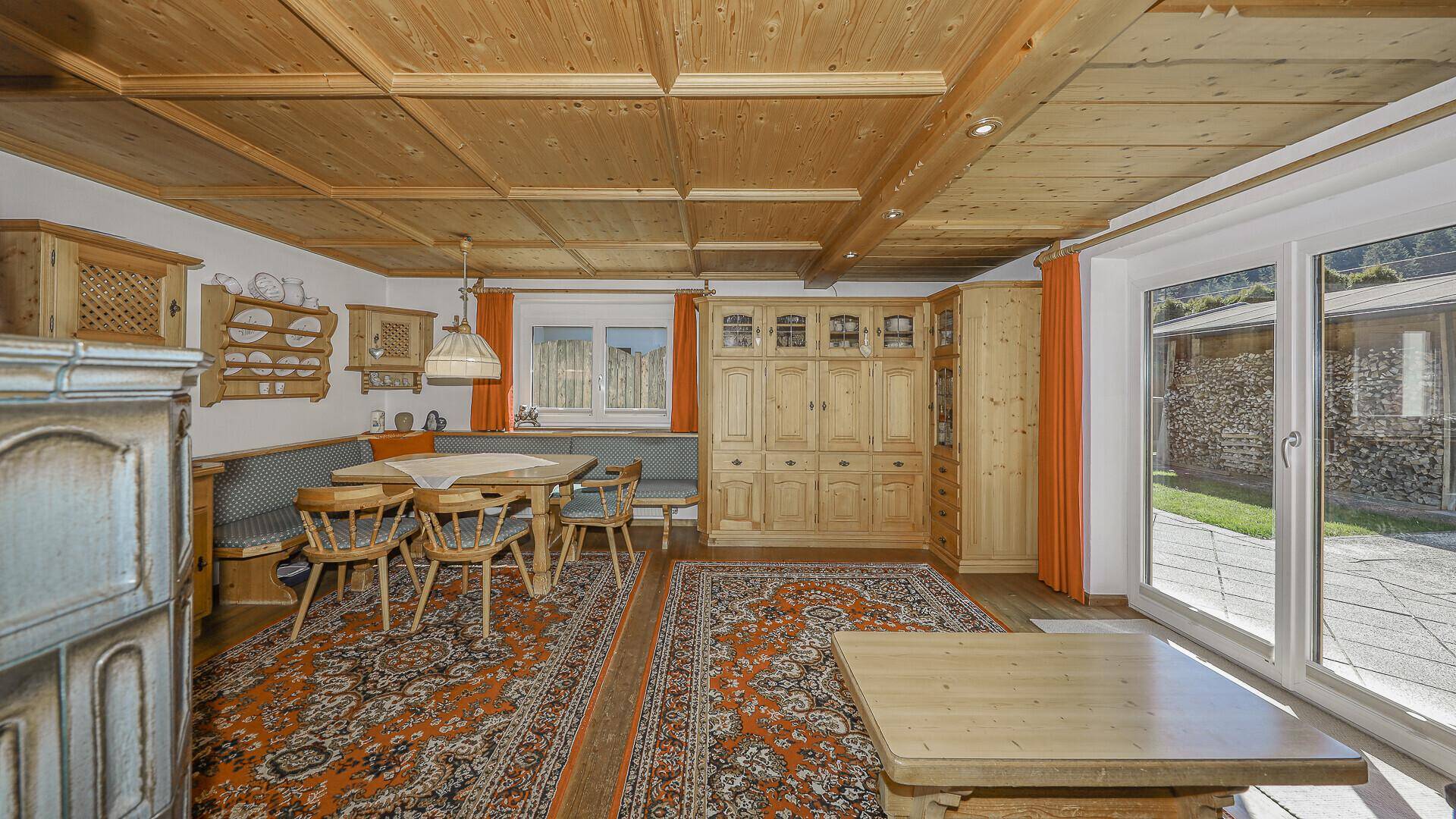 Heller Wohn-Essbereich mit Holztäfelung, Kachelofen und Zugang zum Außenbereich.