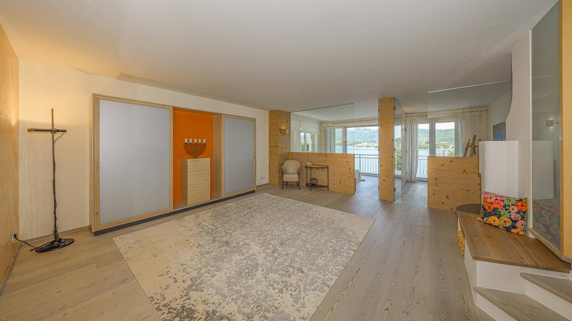Geräumiges Wohnzimmer mit Holzboden, großem Teppich und Zugang zu einem Balkon mit Seeblick.