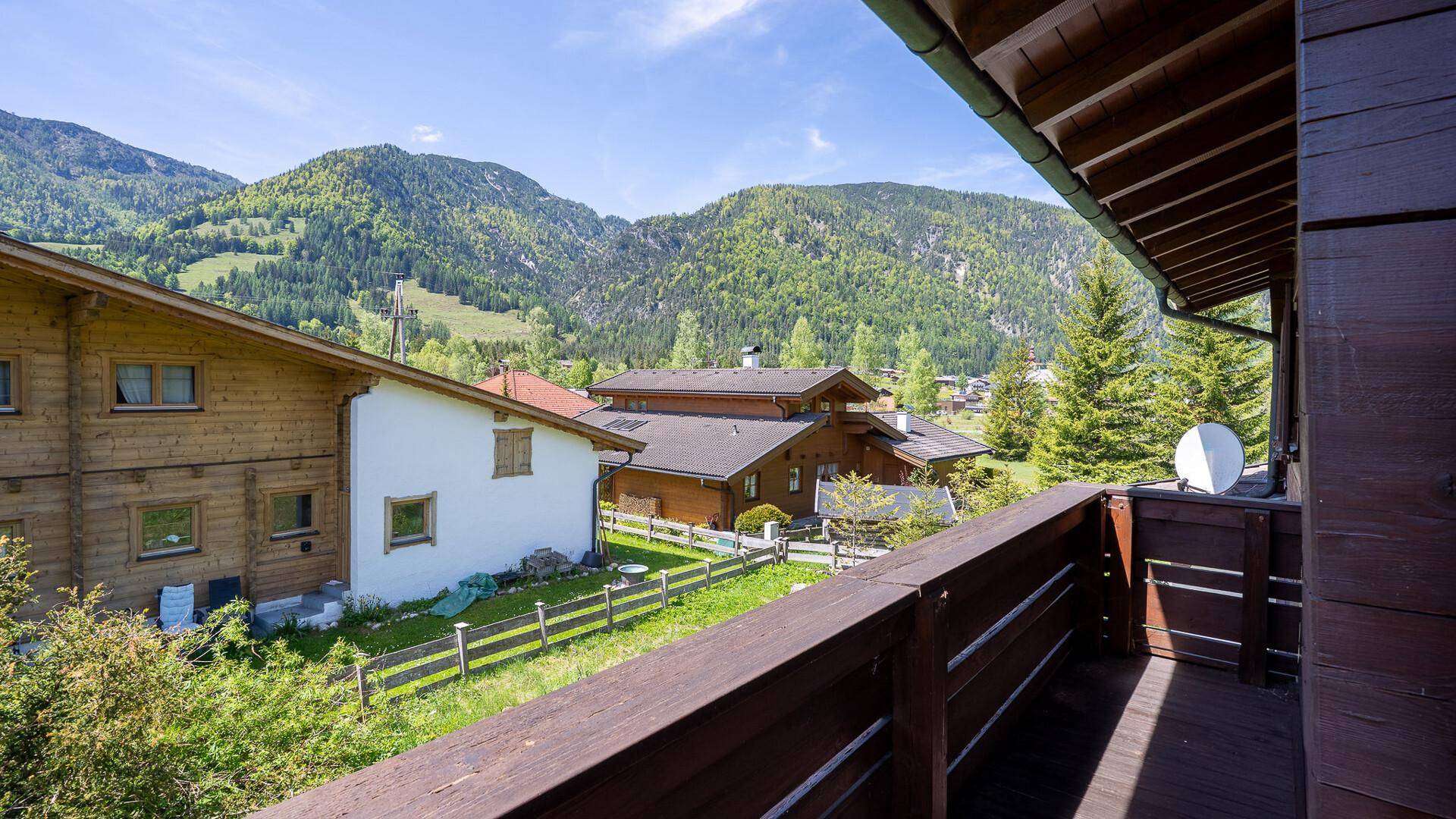 Blick vom Holzbalkon auf die grüne Umgebung, benachbarte Häuser und die beeindruckende Bergkulisse.