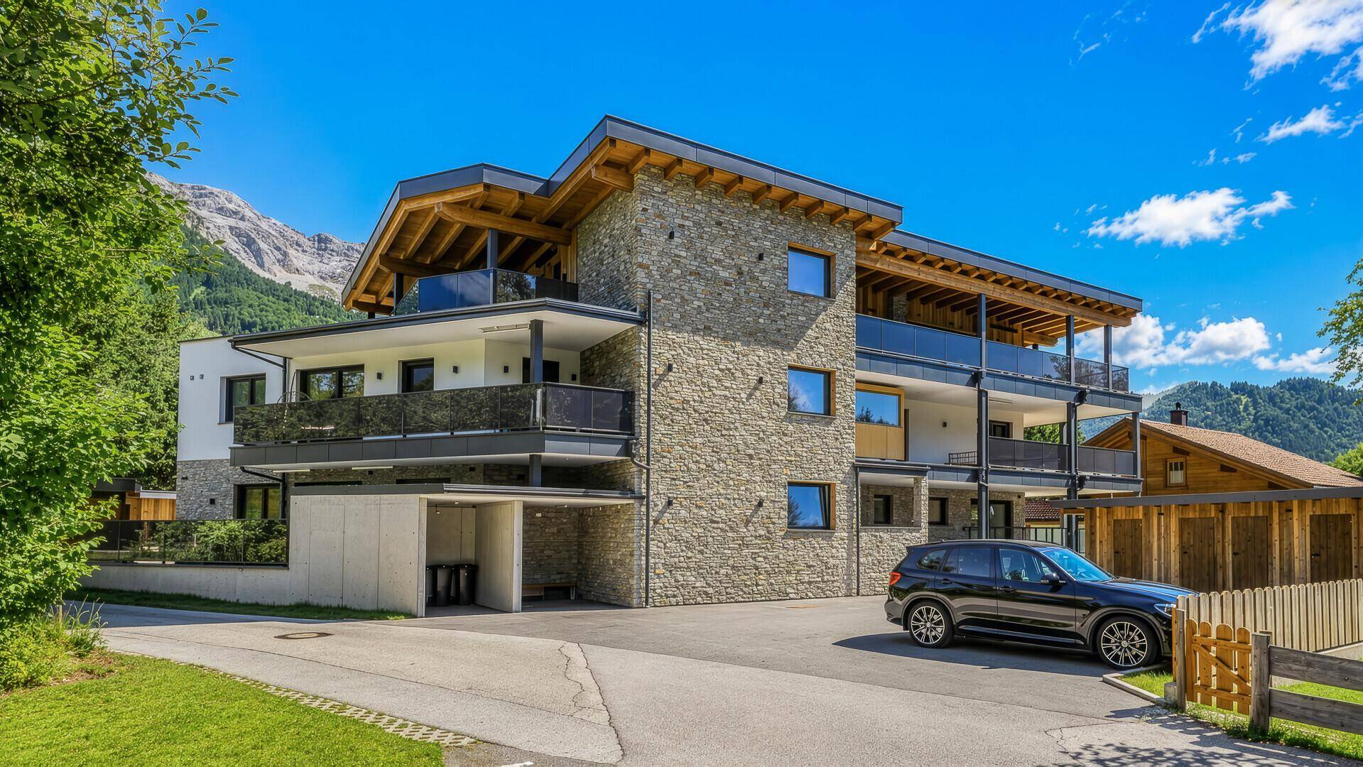 Moderne Gebäudefassade mit Natursteinverkleidung, Parkplatz und Blick auf die umliegende Berglandschaft.