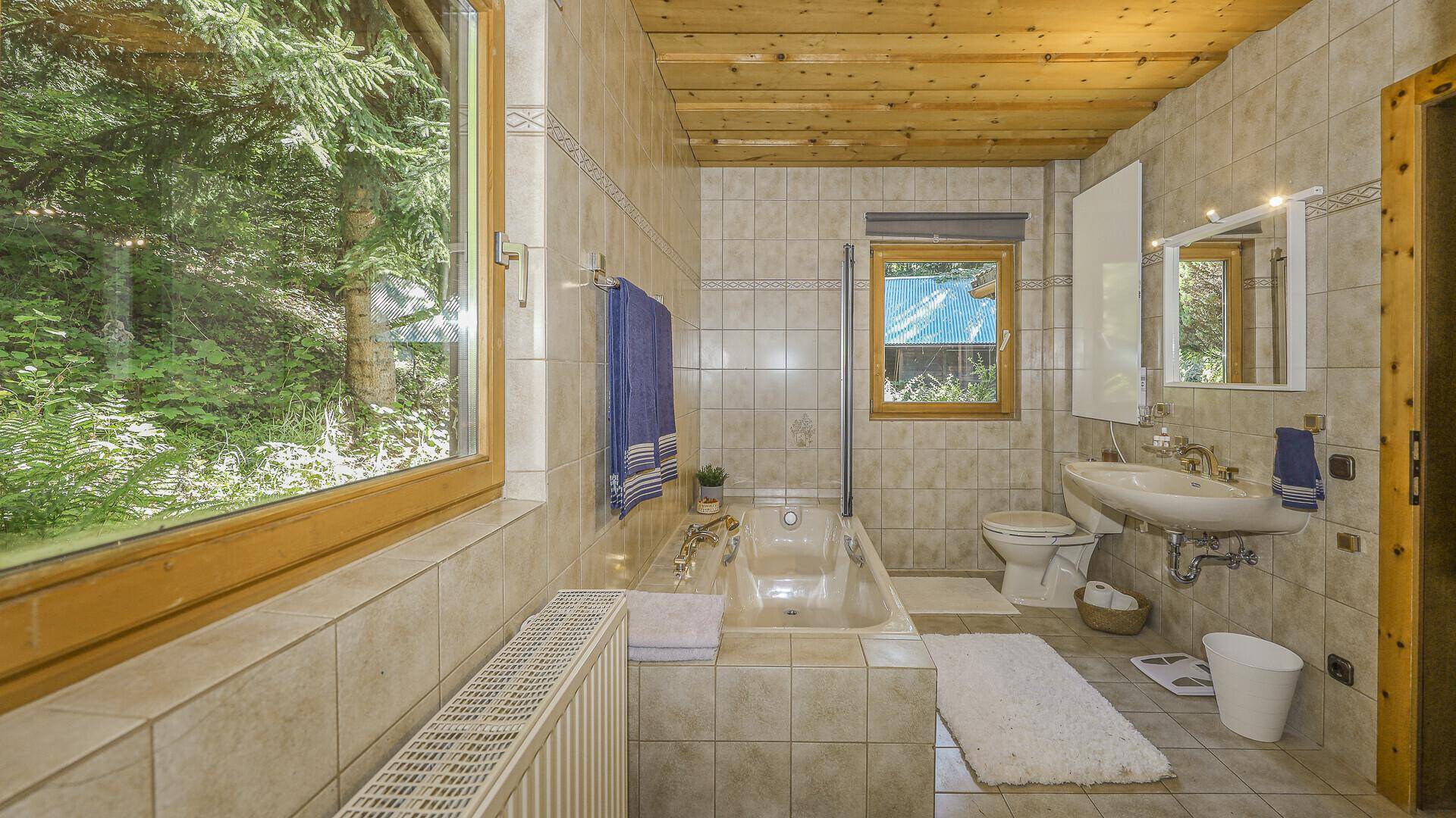 Helles Badezimmer mit Badewanne, Fliesen und Fensterblick in die grüne Natur.