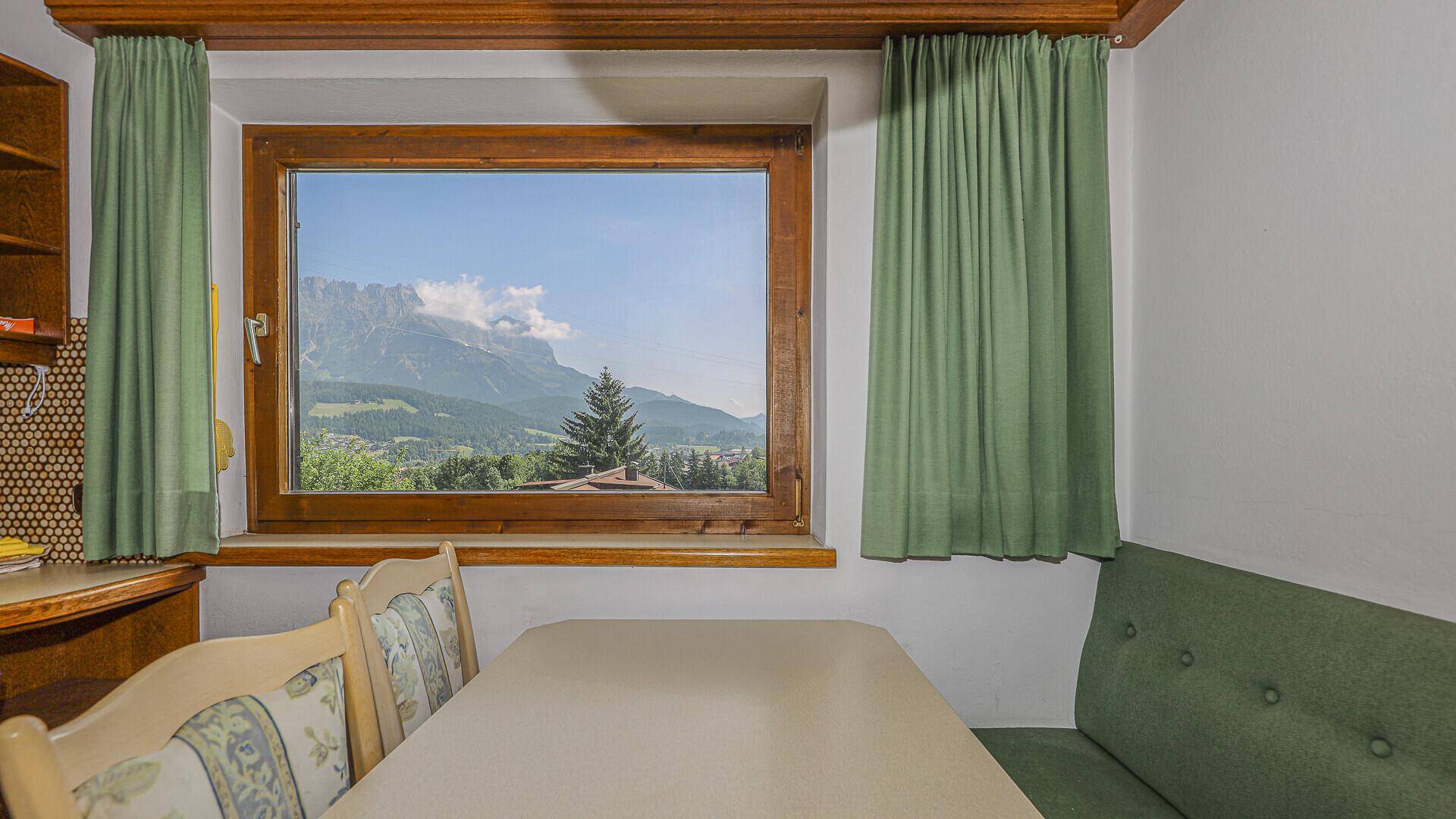 Heller Essbereich mit Holztisch, grünen Vorhängen und weitem Blick auf die Berglandschaft.