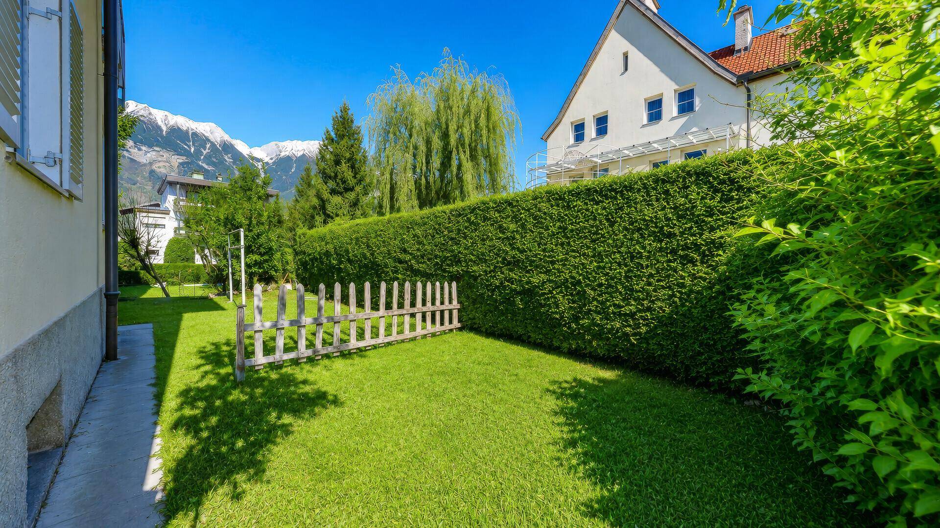 Gepflegter Garten mit Rasenfläche, hoher Hecke und Bergblick im Hintergrund.