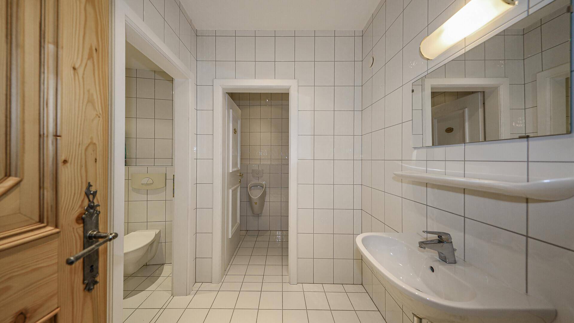 Helles Badezimmer mit weißen Fliesen, Waschbecken und separatem Bereich mit Urinal.