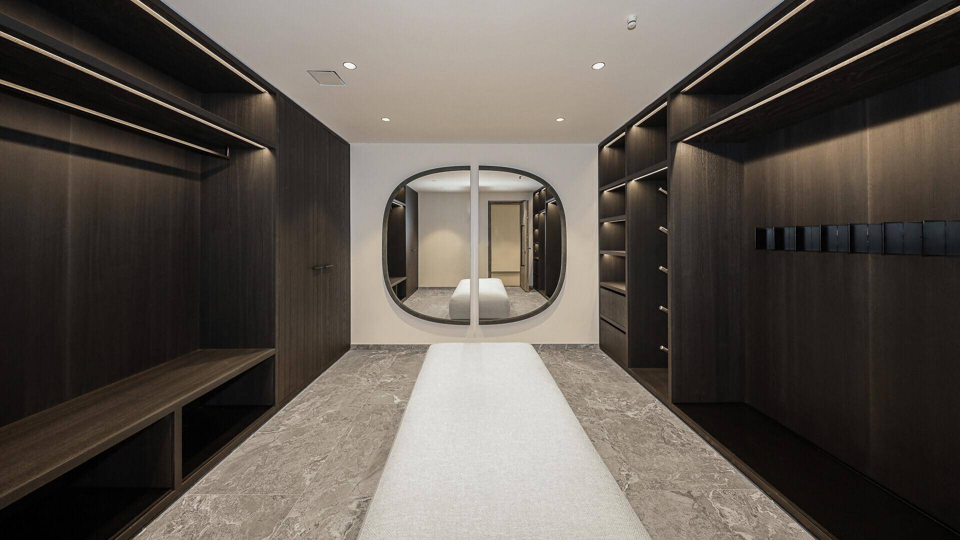 Geräumiger begehbarer Kleiderschrank mit dunklen Holzschränken, zentraler Sitzbank und großem Spiegel.