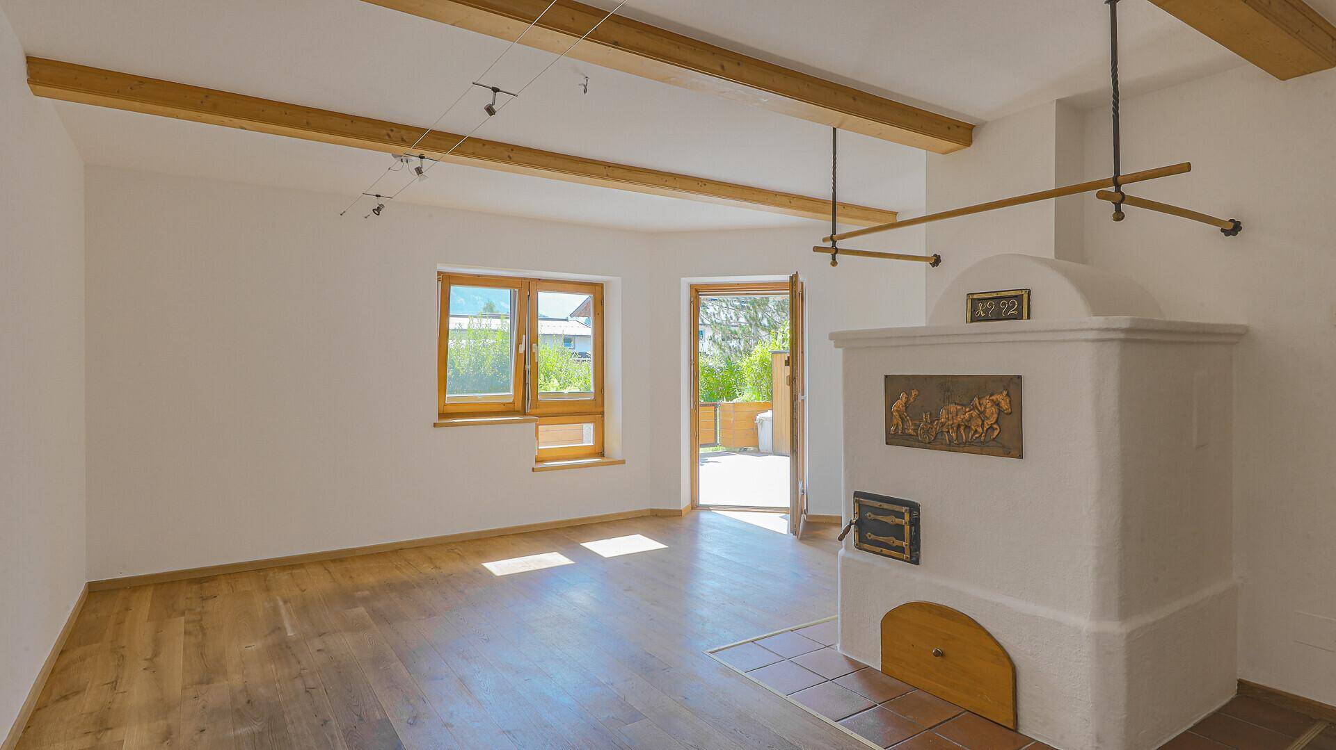 Geräumiges Wohnzimmer mit traditionellem Kachelofen, Holzbalken und Zugang zum Außenbereich.