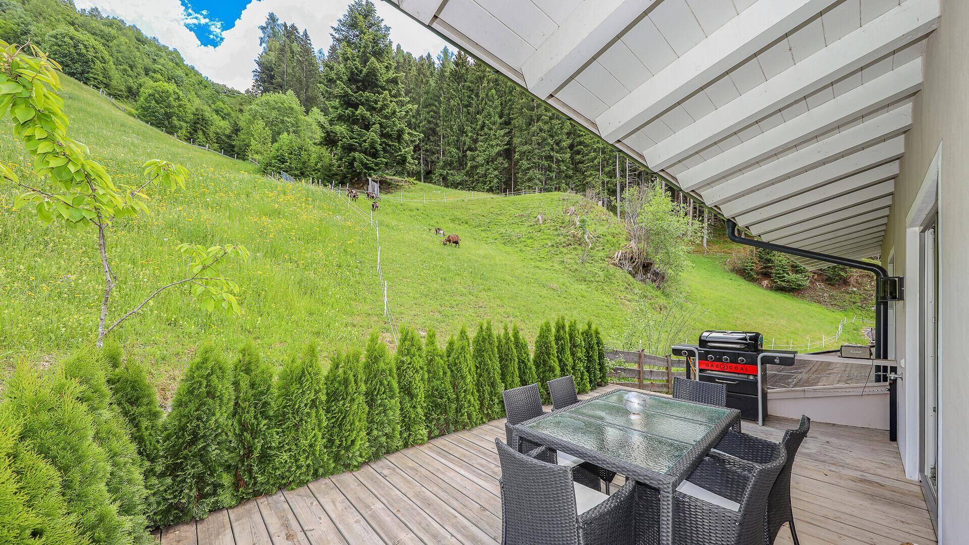 Holzterrasse mit Esstisch und Grill, umgeben von grünen Hecken und Bergblick.