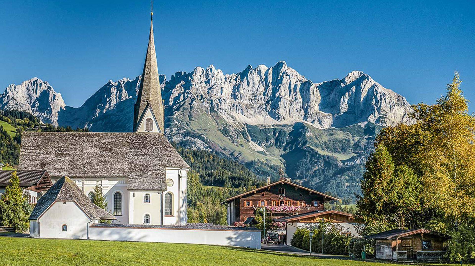 Kirche und traditionelle Häuser vor einer beeindruckenden Bergkulisse an einem sonnigen Sommertag.