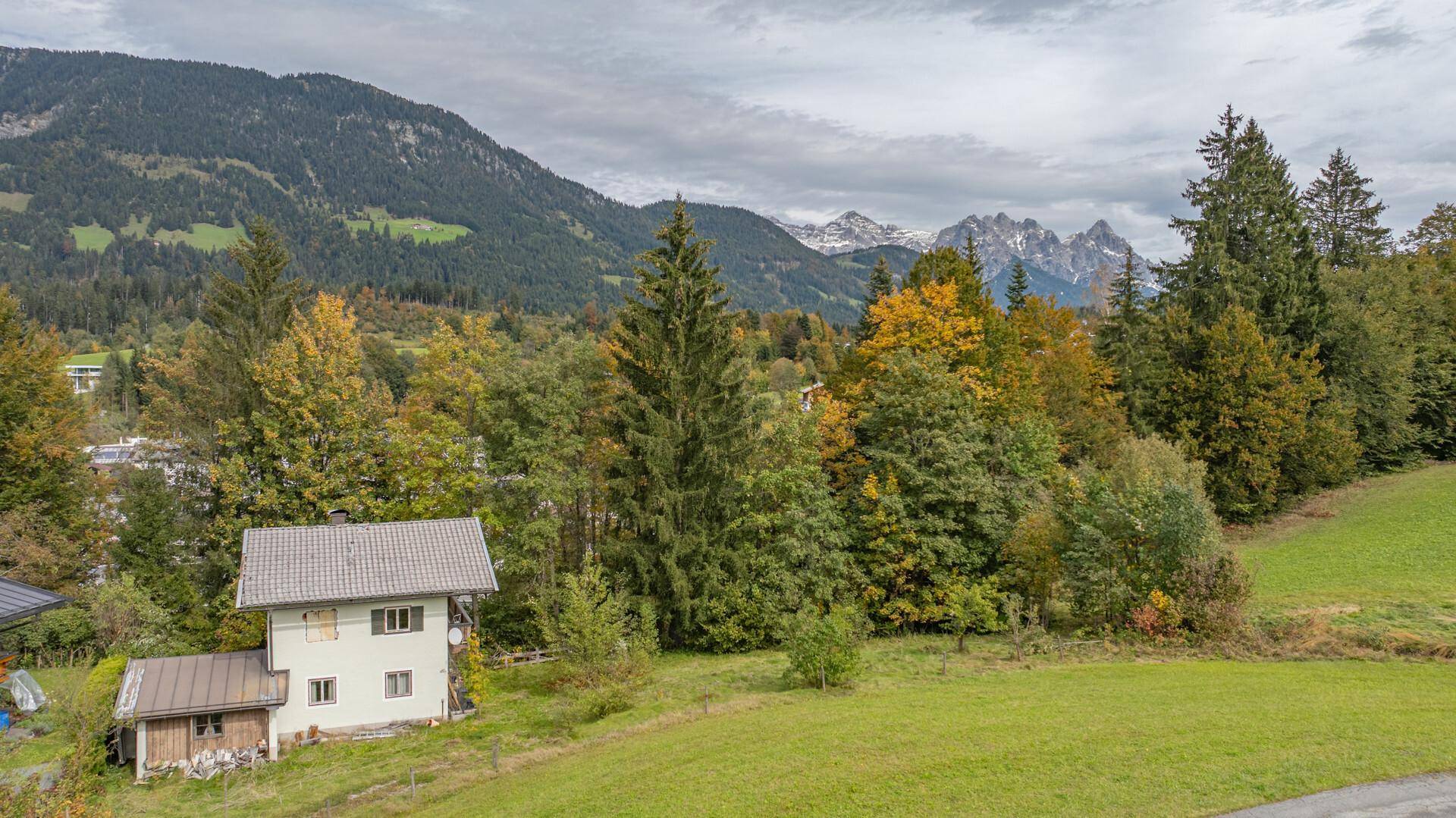 Einzelnes Haus in einer weitläufigen grünen Landschaft, mit bewaldeten Hügeln und schneebedeckten Bergen im Hintergrund.