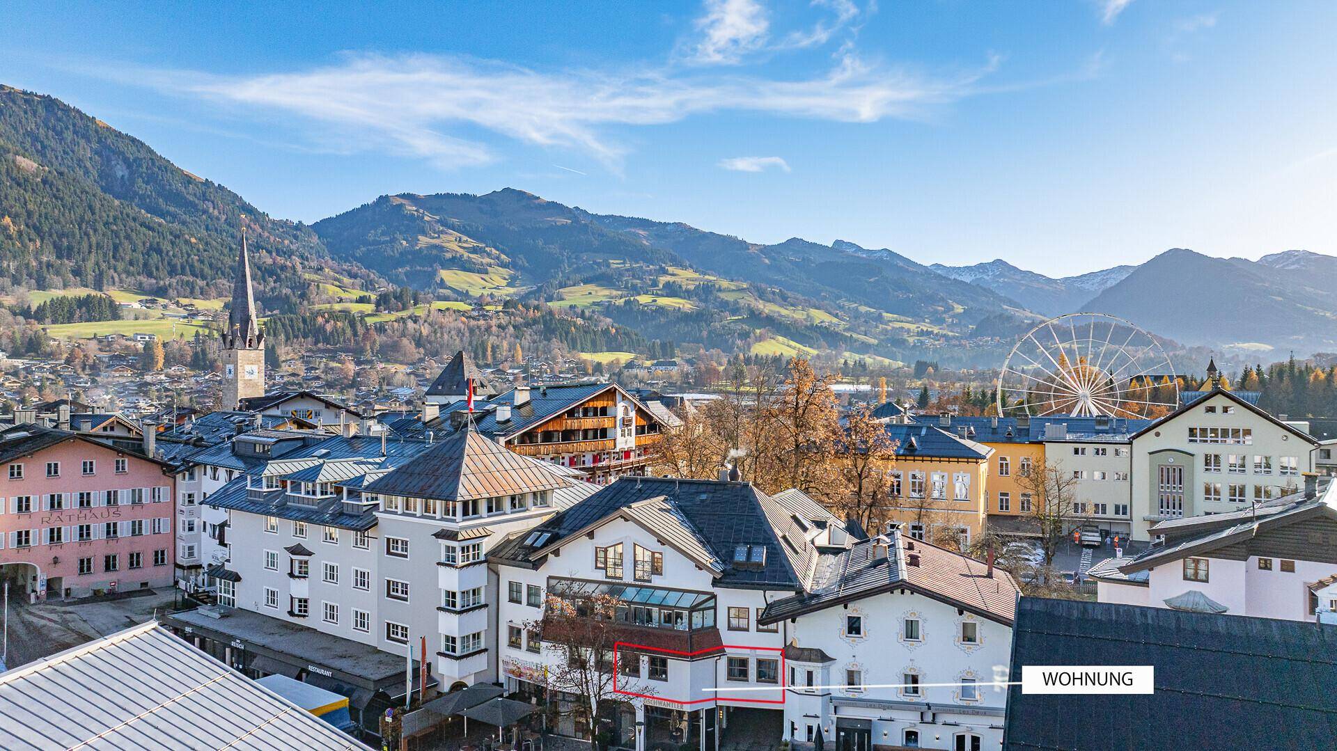 Stadtansicht mit traditionellen Gebäuden, Kirchturm und Riesenrad vor Bergkulisse.