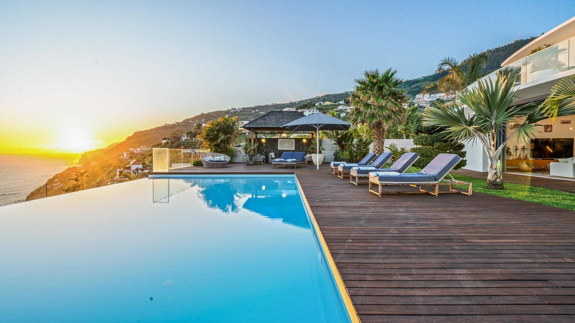 Infinity-Pool mit Holzdeck, Liegestühlen und Panoramablick auf das Meer bei Sonnenuntergang.