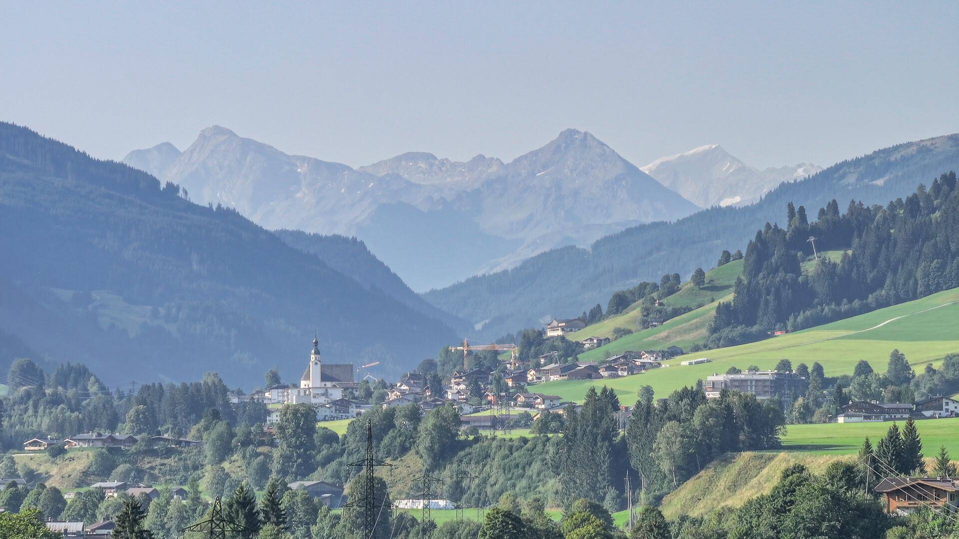 Panoramablick auf ein malerisches Dorf in einem grünen Tal, umgeben von majestätischen Bergen.