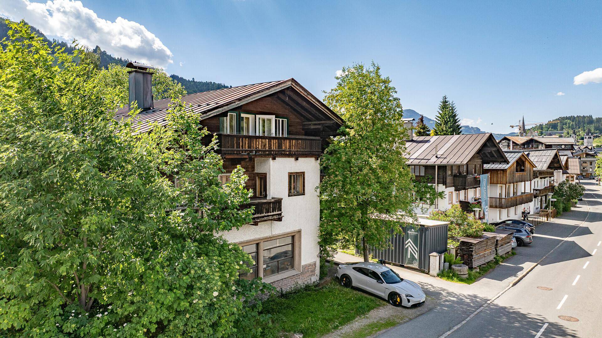 Traditionelles Haus mit Holzbalkonen und weißer Fassade, umgeben von Bäumen an einer Straße.
