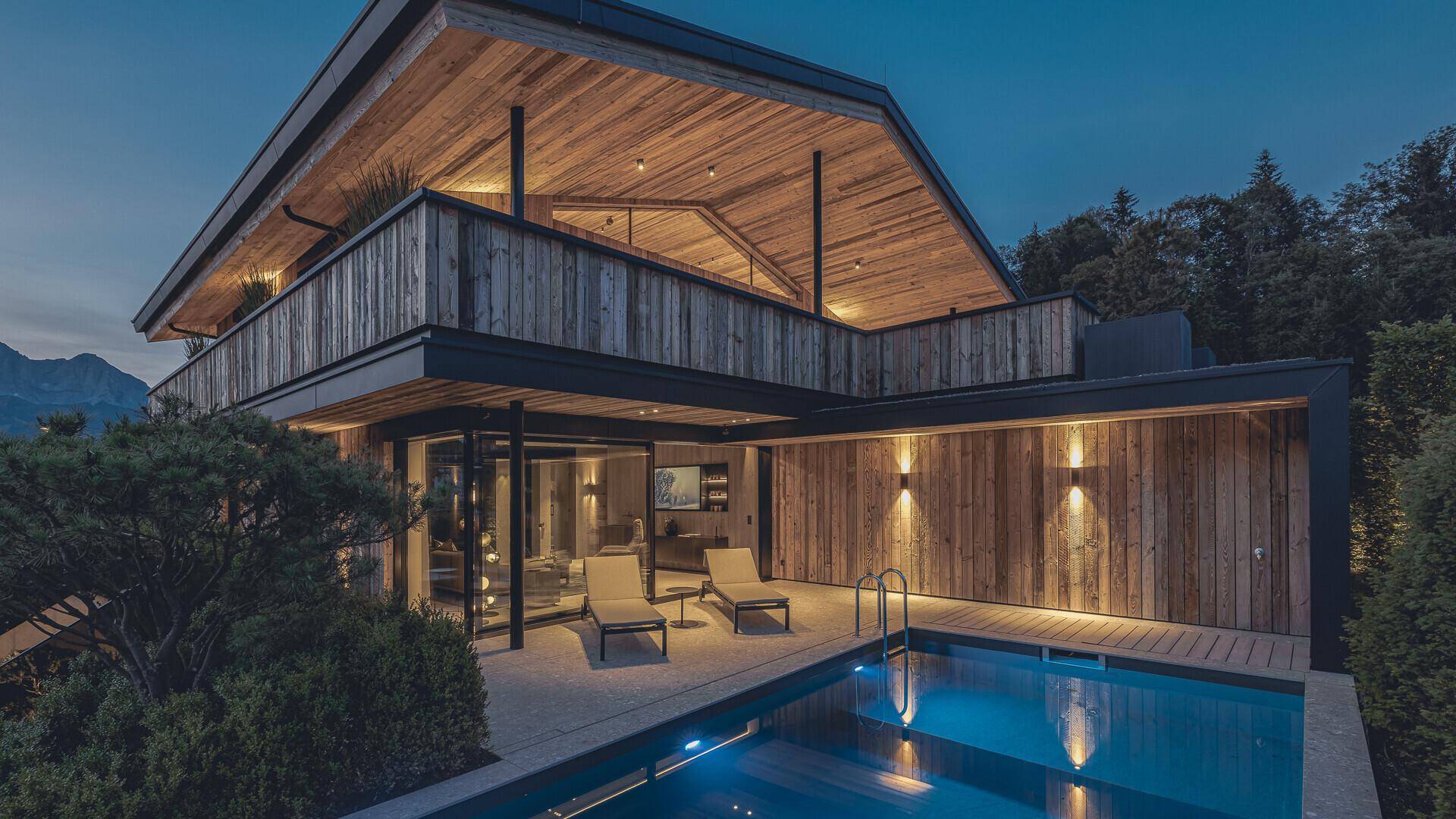 Elegantes Holzhaus bei Nacht mit beleuchtetem Pool und gemütlicher Terrasse.