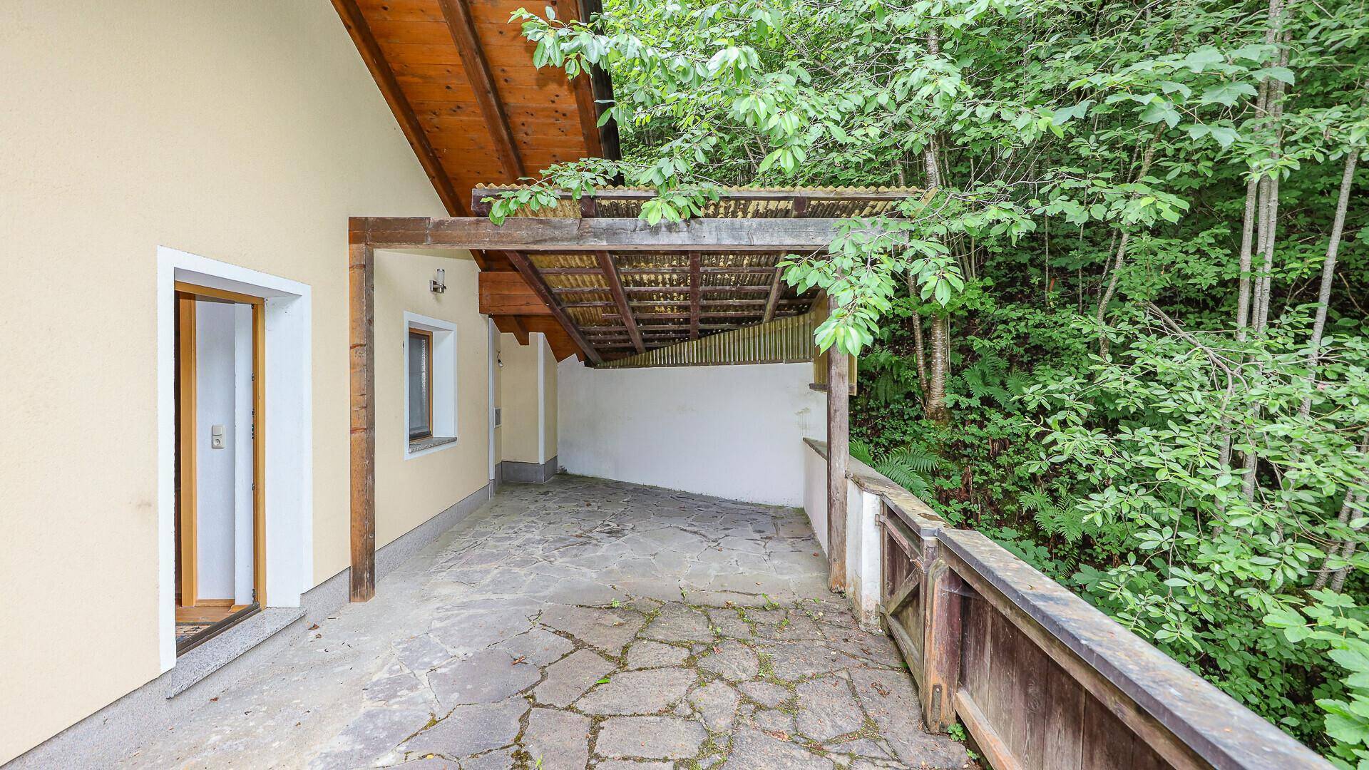 Überdachte Terrasse mit Steinboden und direktem Zugang zum bewaldeten Gartenbereich.