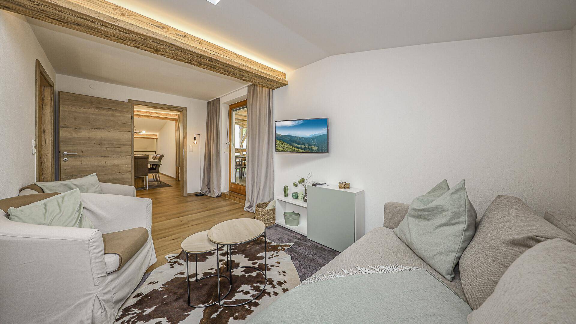 Gemütliches Wohnzimmer mit Sofa, Fernseher und Zugang zu einem Balkon oder einer Terrasse, unter Holzbalken.