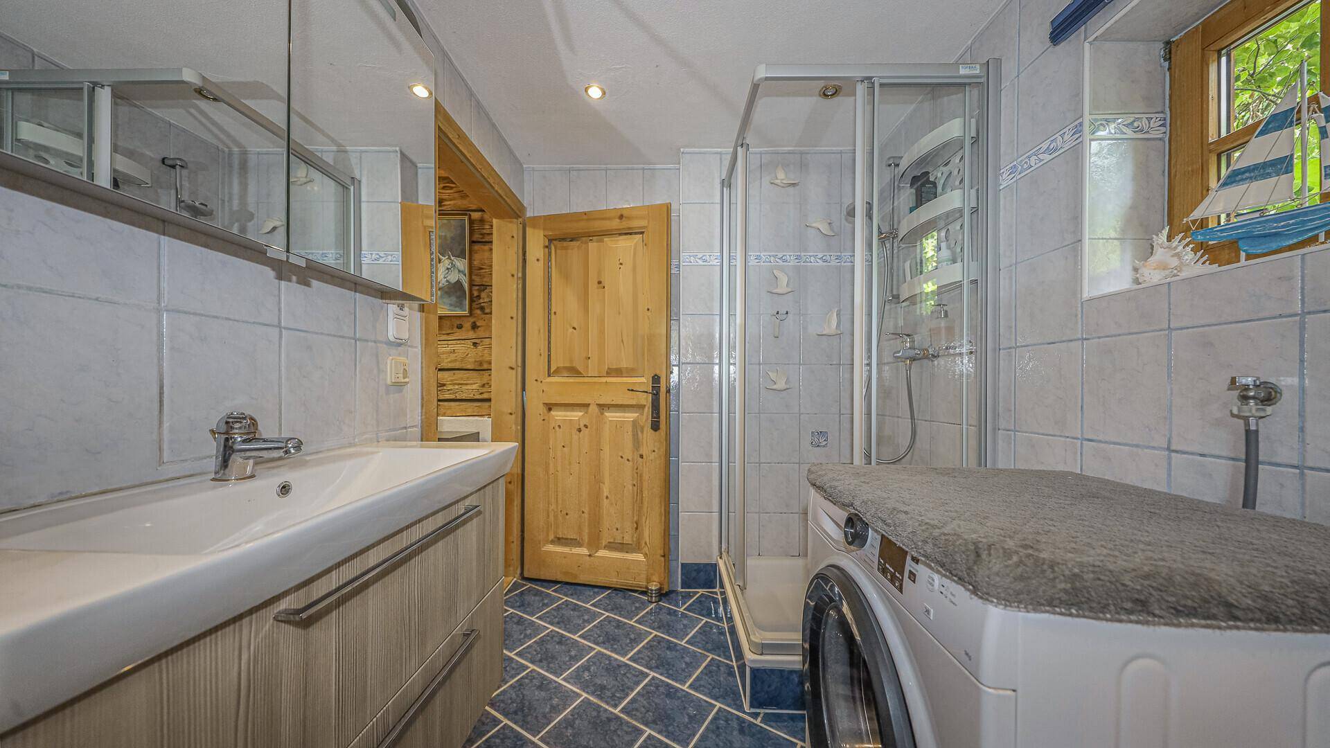 Badezimmer mit Dusche, Waschbecken, Spiegel und Waschmaschine, gefliest in Blau und Weiß.