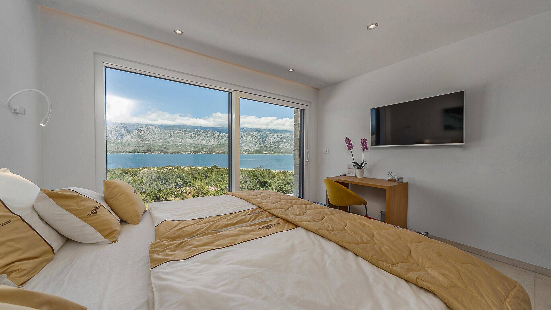 Komfortables Schlafzimmer mit Doppelbett, Wandfernseher und raumhohen Fenstern mit Panoramablick auf das Meer.