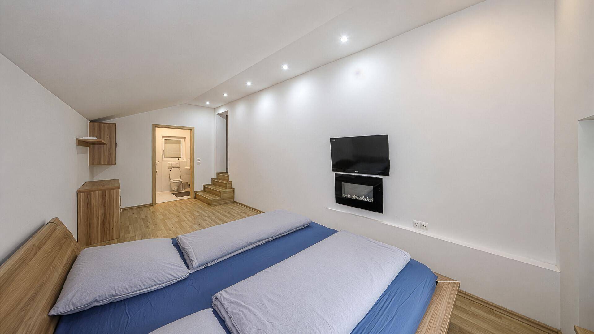 Modern eingerichtetes Schlafzimmer mit Holzboden, Wandfernseher und Zugang zu einem Badezimmer.