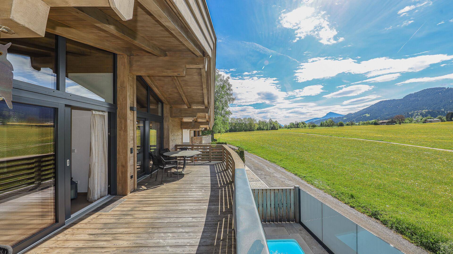 Großer Holzbalkon mit Sitzgelegenheiten und weitem Blick über grüne Felder und Berge.