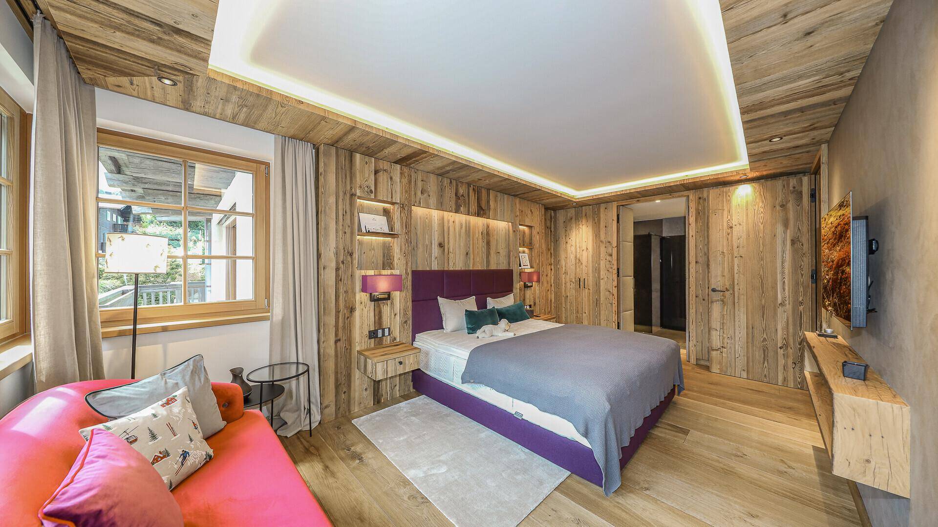 Helles Schlafzimmer mit Holzwänden, lila Bett und einem bequemen Sofa am Fenster.