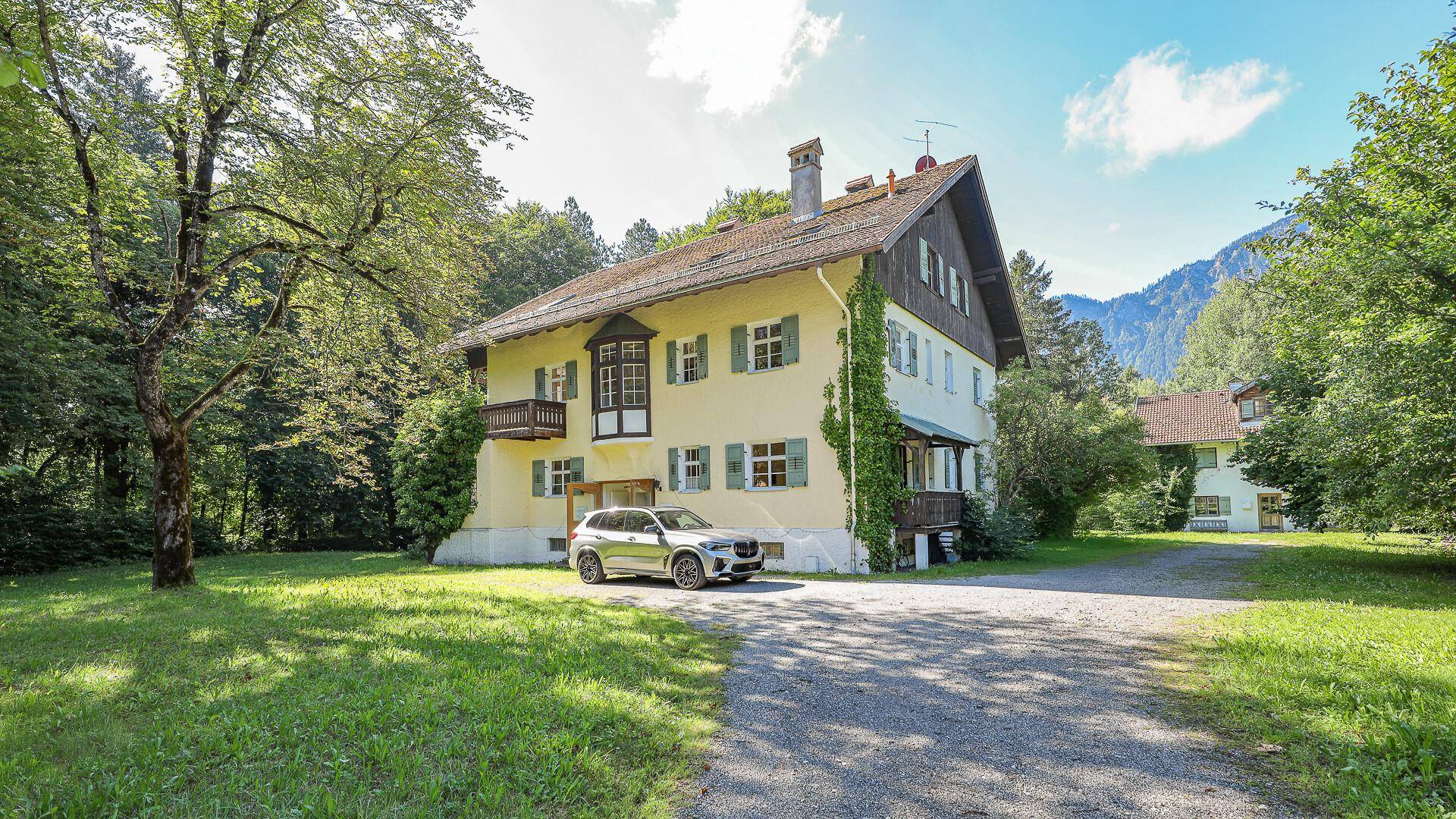 Hausansicht mit Kiesauffahrt und geparktem Auto, umgeben von Bäumen und Bergszenerie.