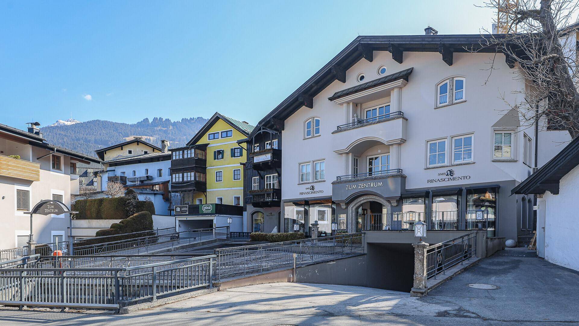 Gebäude im alpinen Stil mit Parkplatz und Zufahrt zur Tiefgarage, umgeben von Bergen.