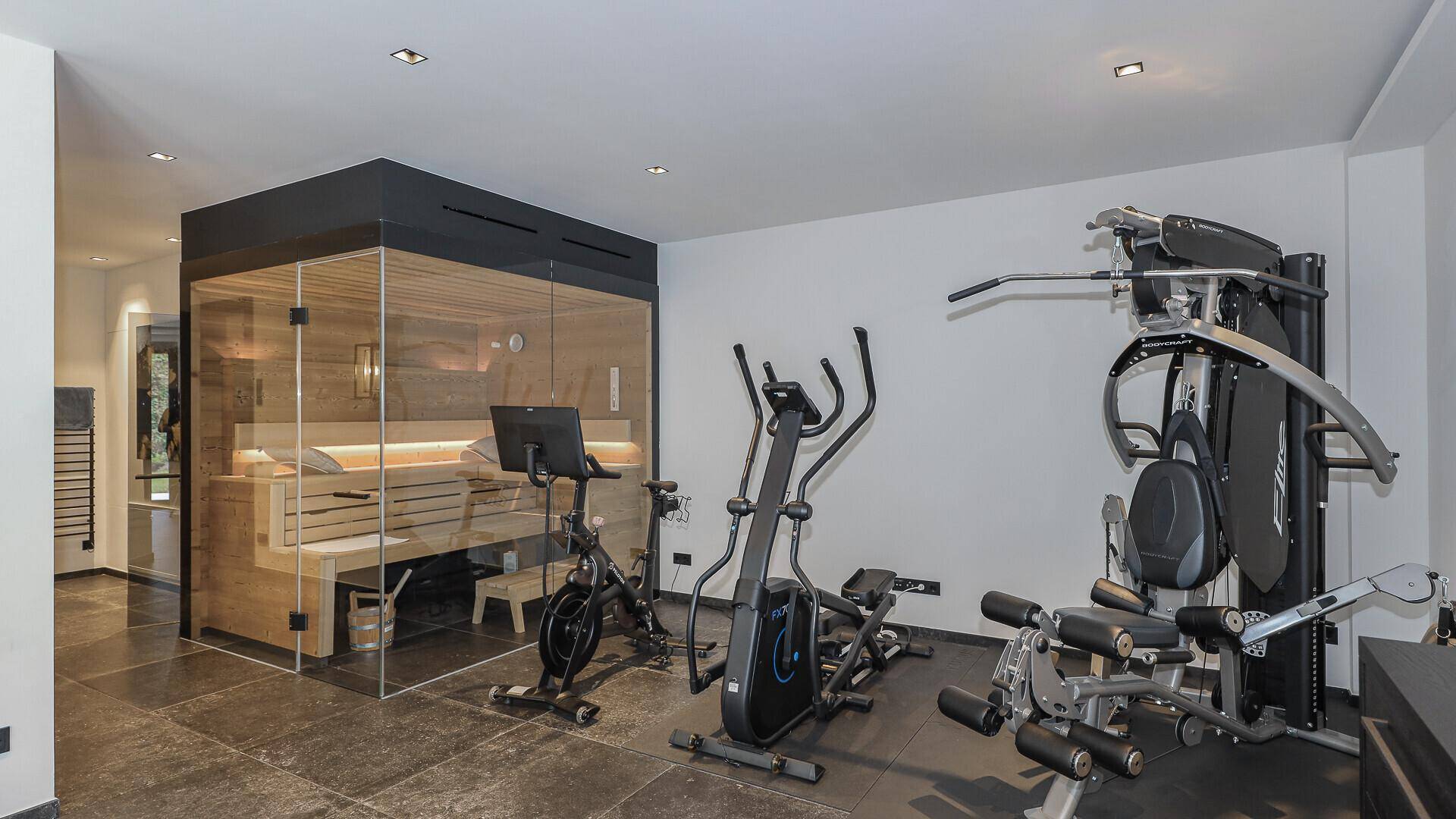 Moderner Fitnessbereich mit Trainingsgeräten und einer stilvollen Glas-Saunakabine.