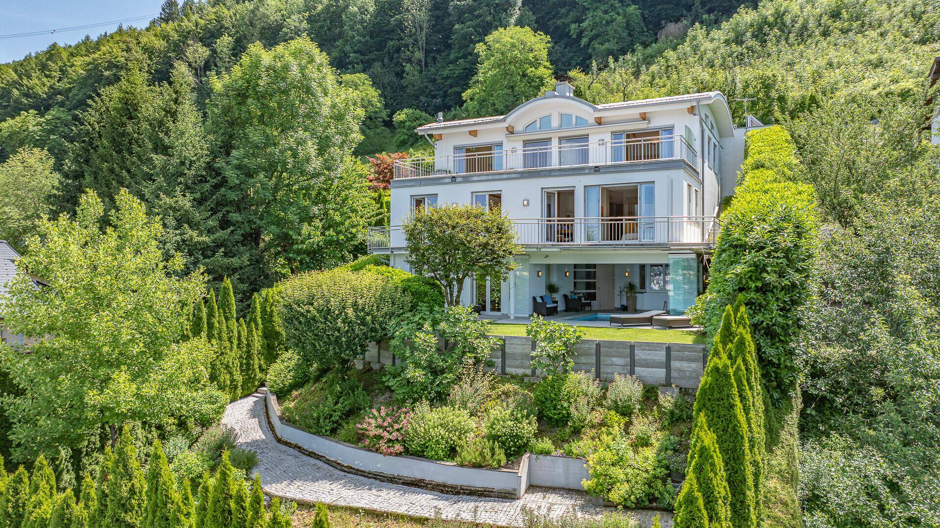 Elegantes Haus an einem Hang, umgeben von üppigem Grün und gepflegten Gärten mit altem Baumbestand.