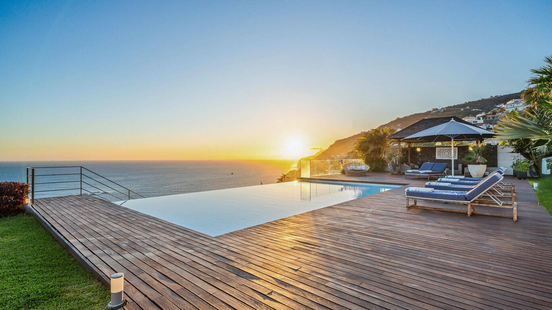 Malerischer Poolbereich bei Sonnenuntergang mit Holzdeck, Sonnenliegen und schattigem Pavillon, direkt am Meer.