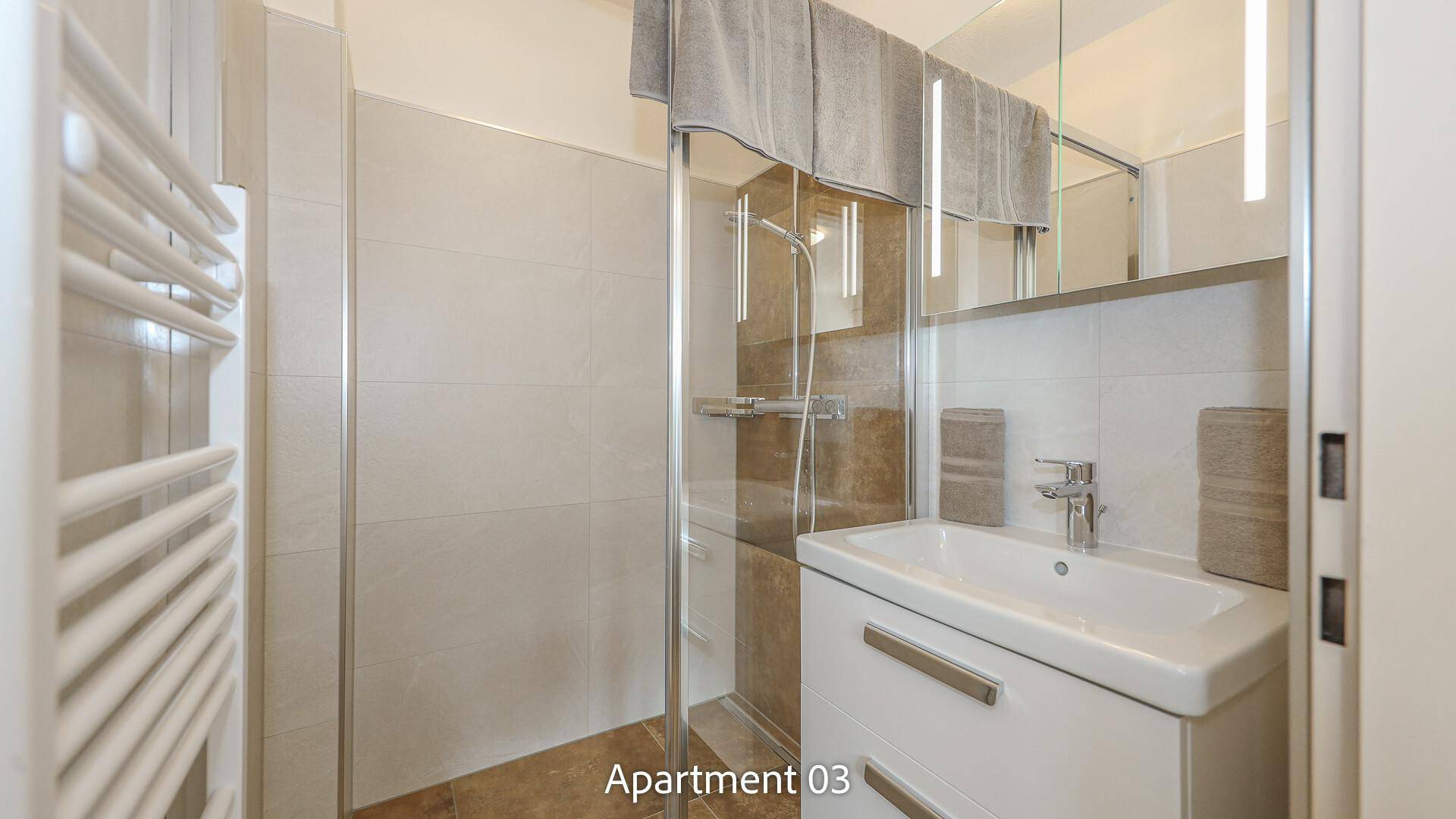 Modernes Badezimmer von Apartment 03 mit hellen Fliesen, Waschtisch, WC und einer geräumigen Duschkabine.