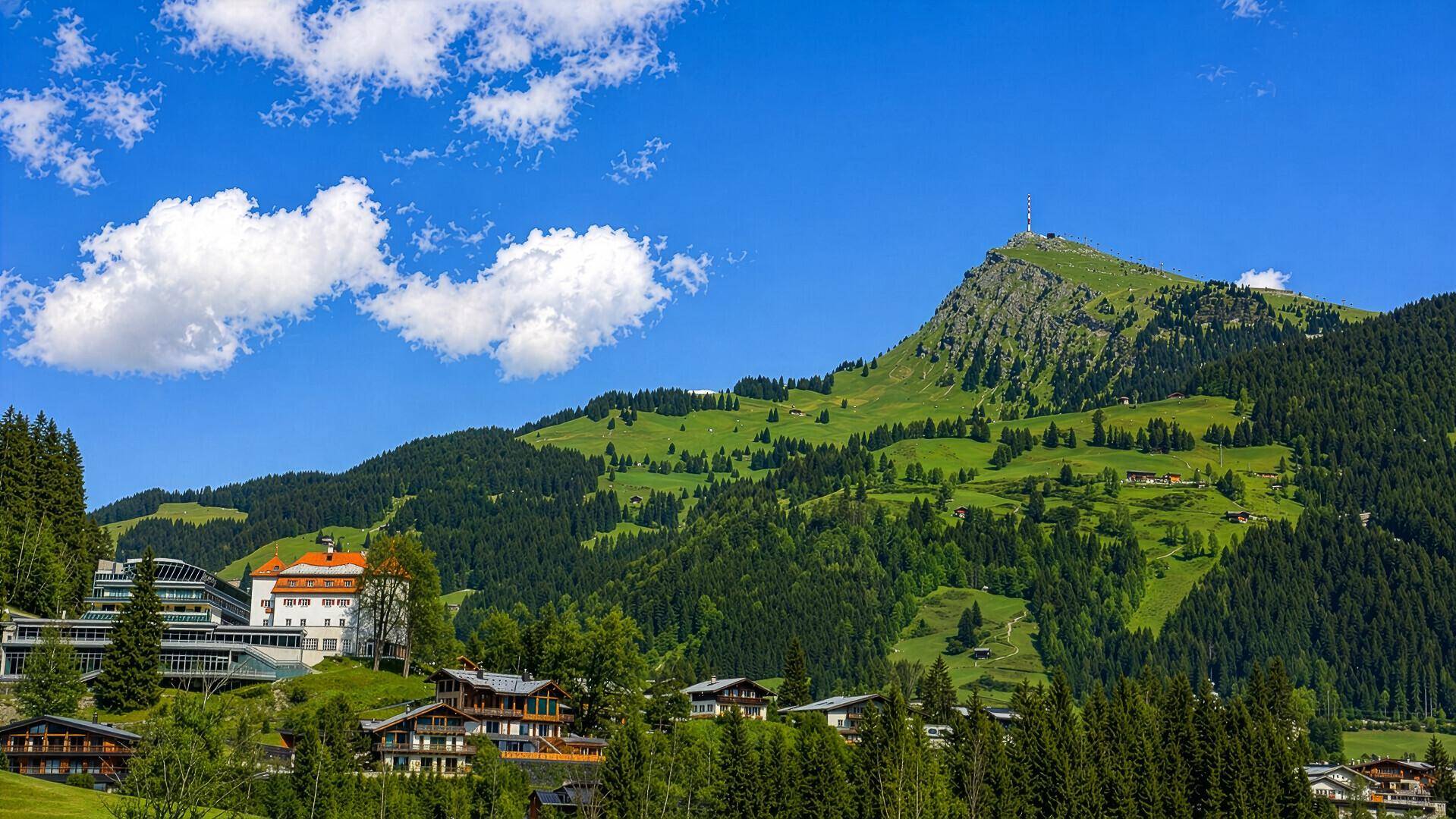 Panoramablick auf die grüne Berglandschaft mit vereinzelten Häusern unter blauem Himmel.