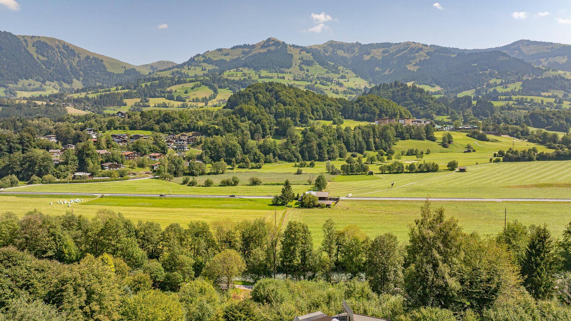 Weitläufige grüne Landschaft mit Feldern, Wäldern und sanften Hügeln unter blauem Himmel.