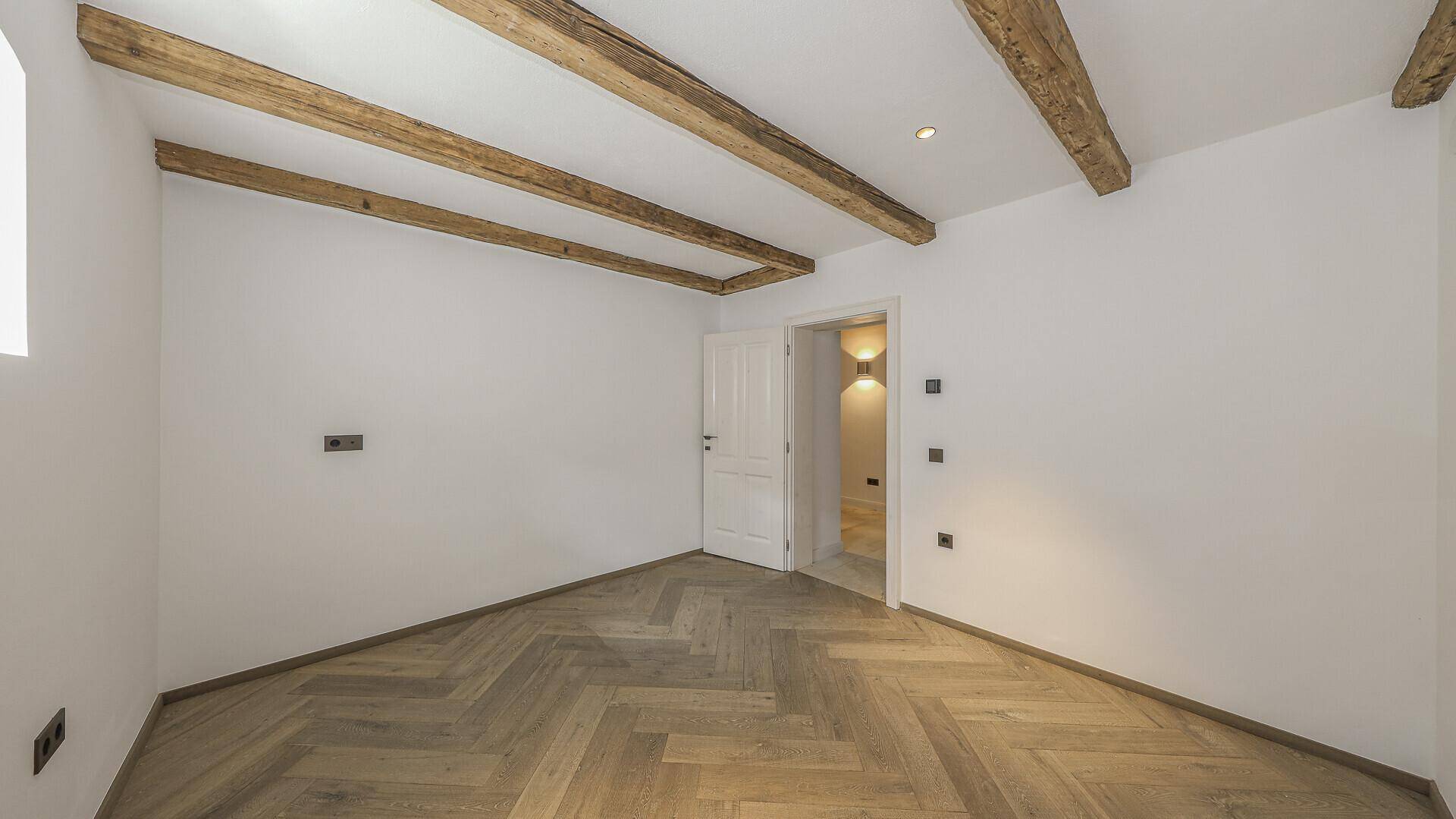 Geräumiges Zimmer mit Fischgrätparkett, weißen Wänden und Holzbalkendecke.