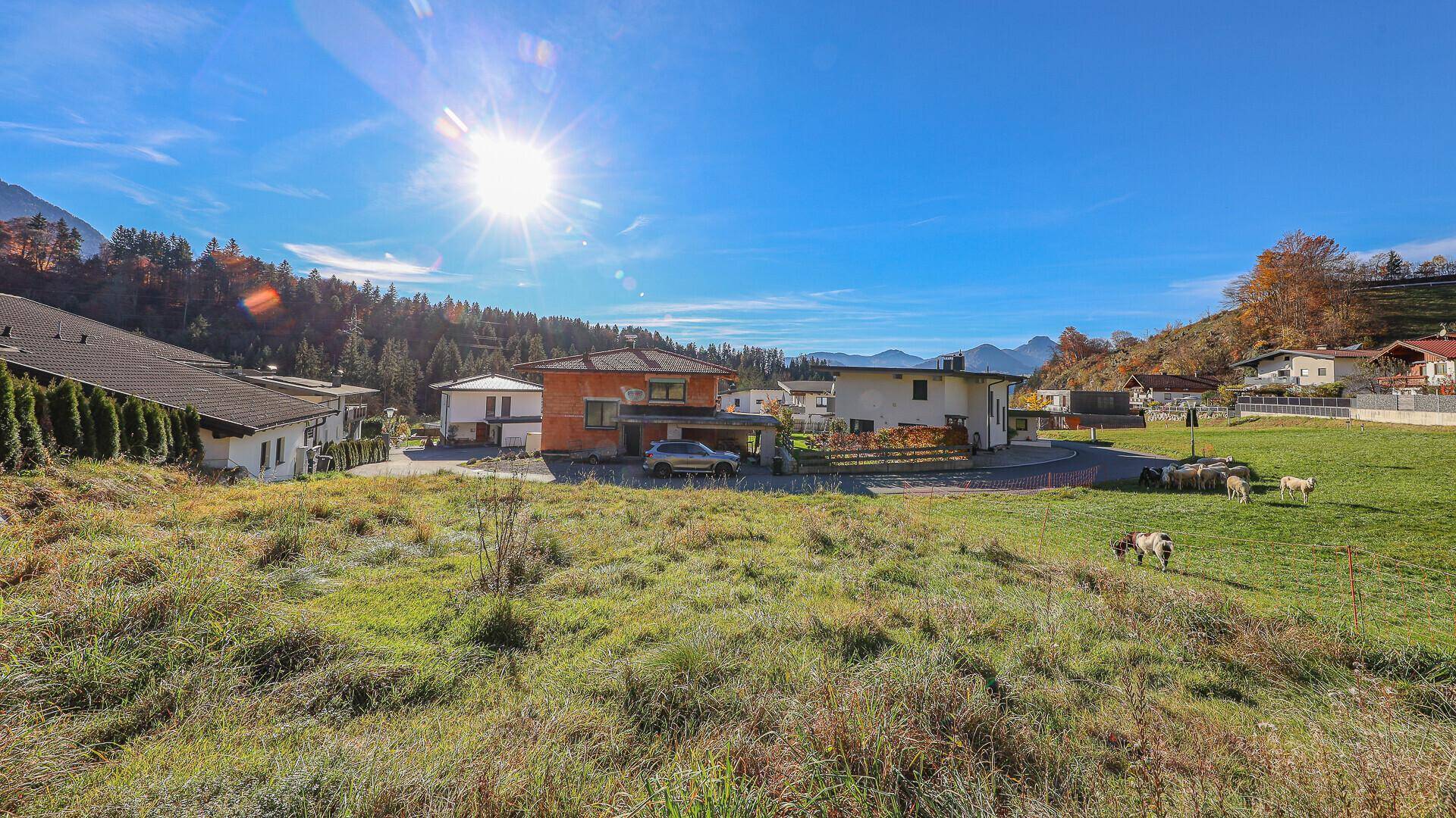 Weitläufige grüne Landschaft mit Häusern und einem beeindruckenden Bergpanorama bei Sonnenschein.