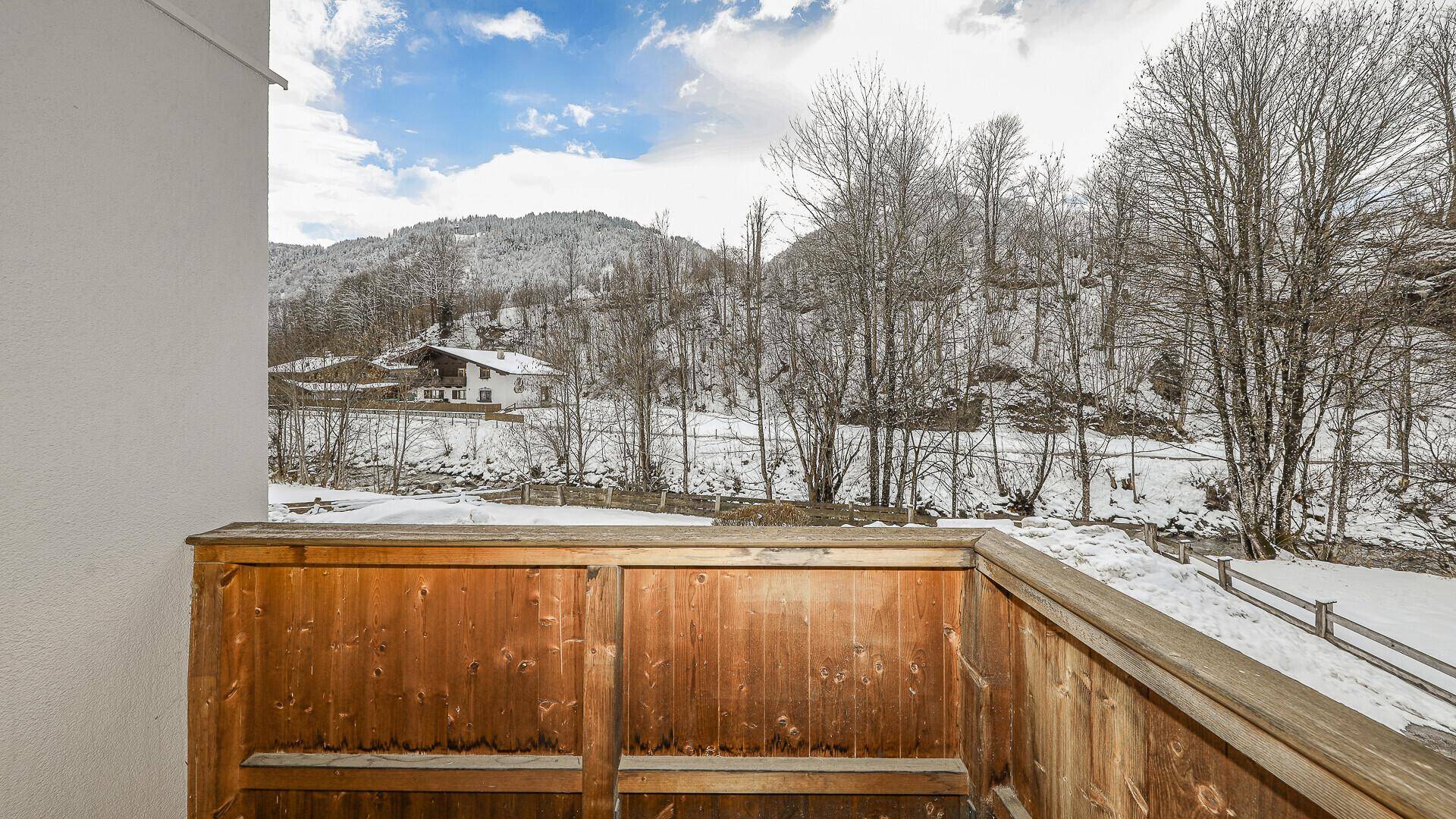 Balkon mit Holzbalkonbrüstung und Blick auf eine verschneite Berglandschaft mit Bäumen und Häusern.