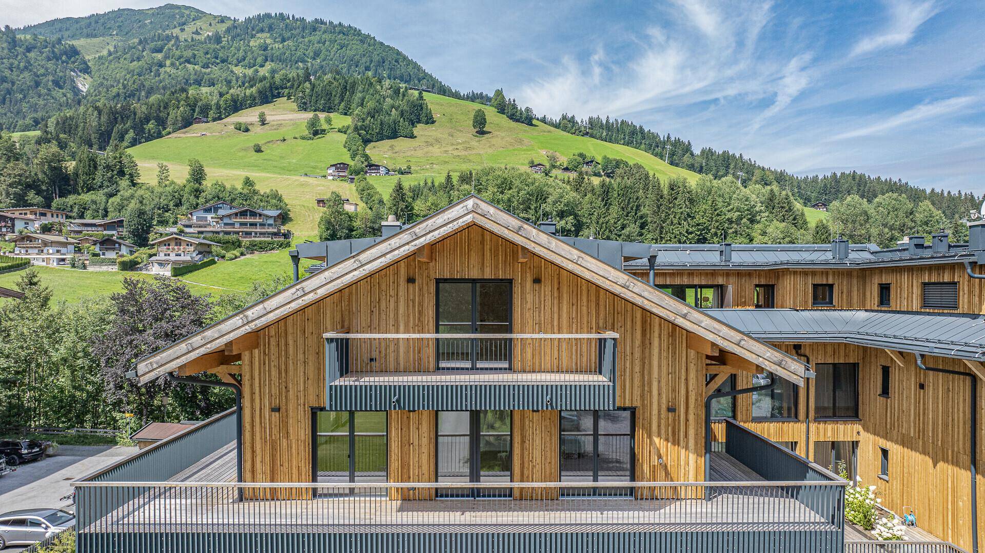 Modernes Apartmentgebäude im alpinen Stil mit Holzfassade und Balkonen vor grüner Bergkulisse.