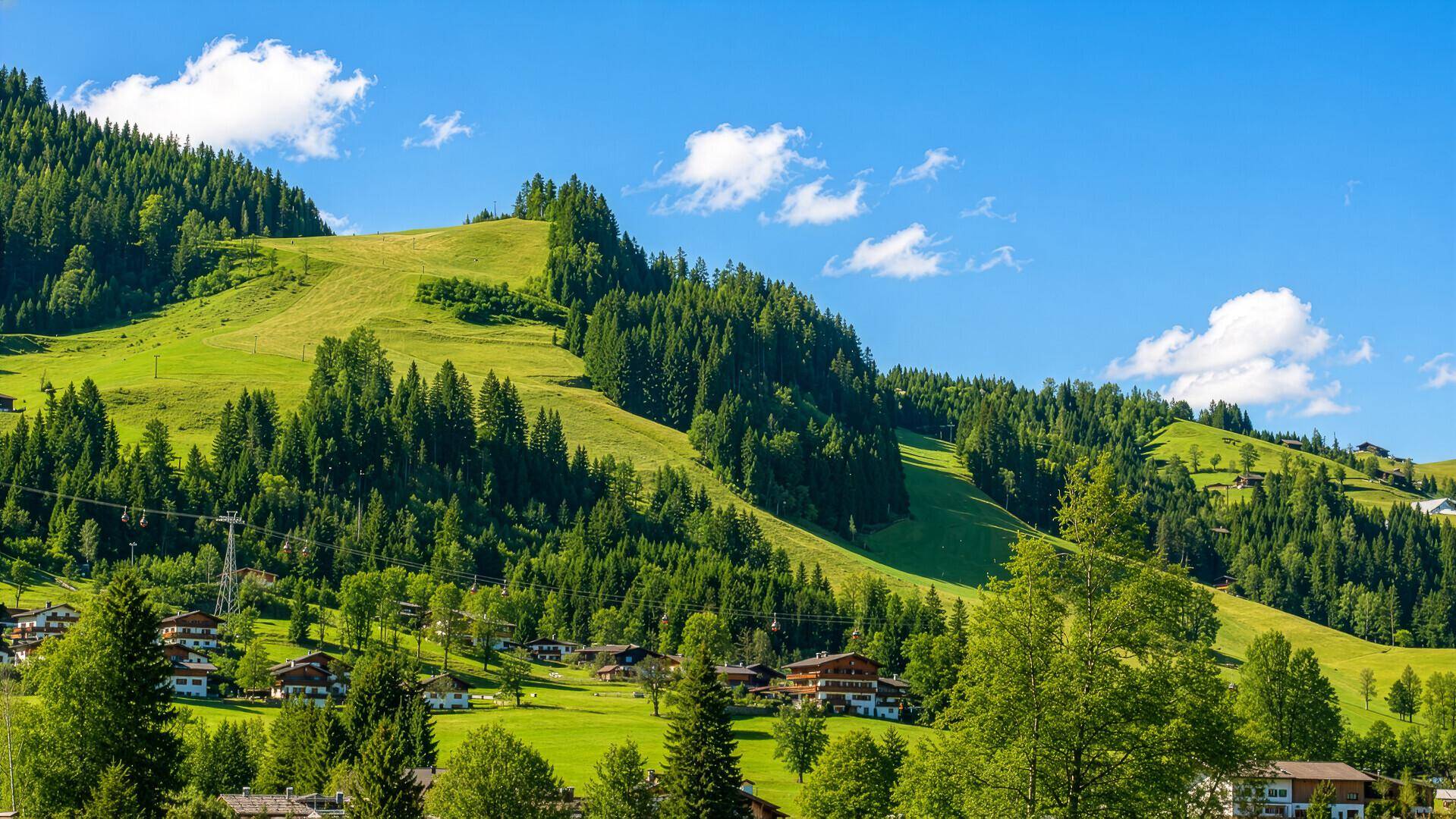 Weitläufige grüne Berglandschaft mit verstreuten Häusern und dichtem Wald unter blauem Himmel.