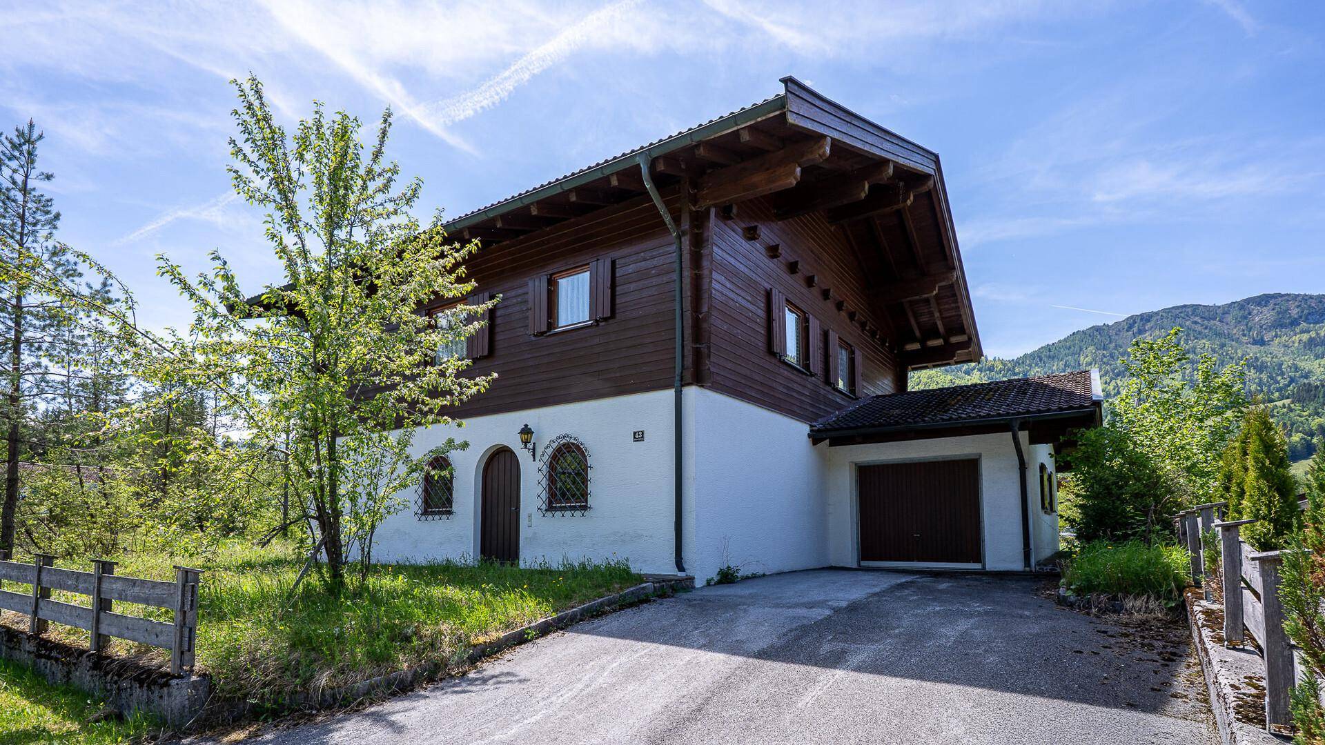 Traditionelles Chalet mit integrierter Garage und Zufahrt, umgeben von Bäumen und Bergen.