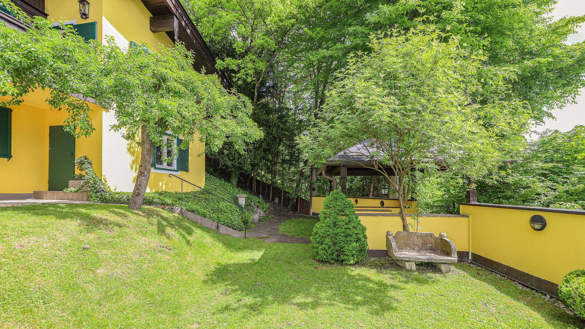 Idyllischer Garten mit Rasen, Steinbank, Gartenlaube und Zugang zum gelben Haus.