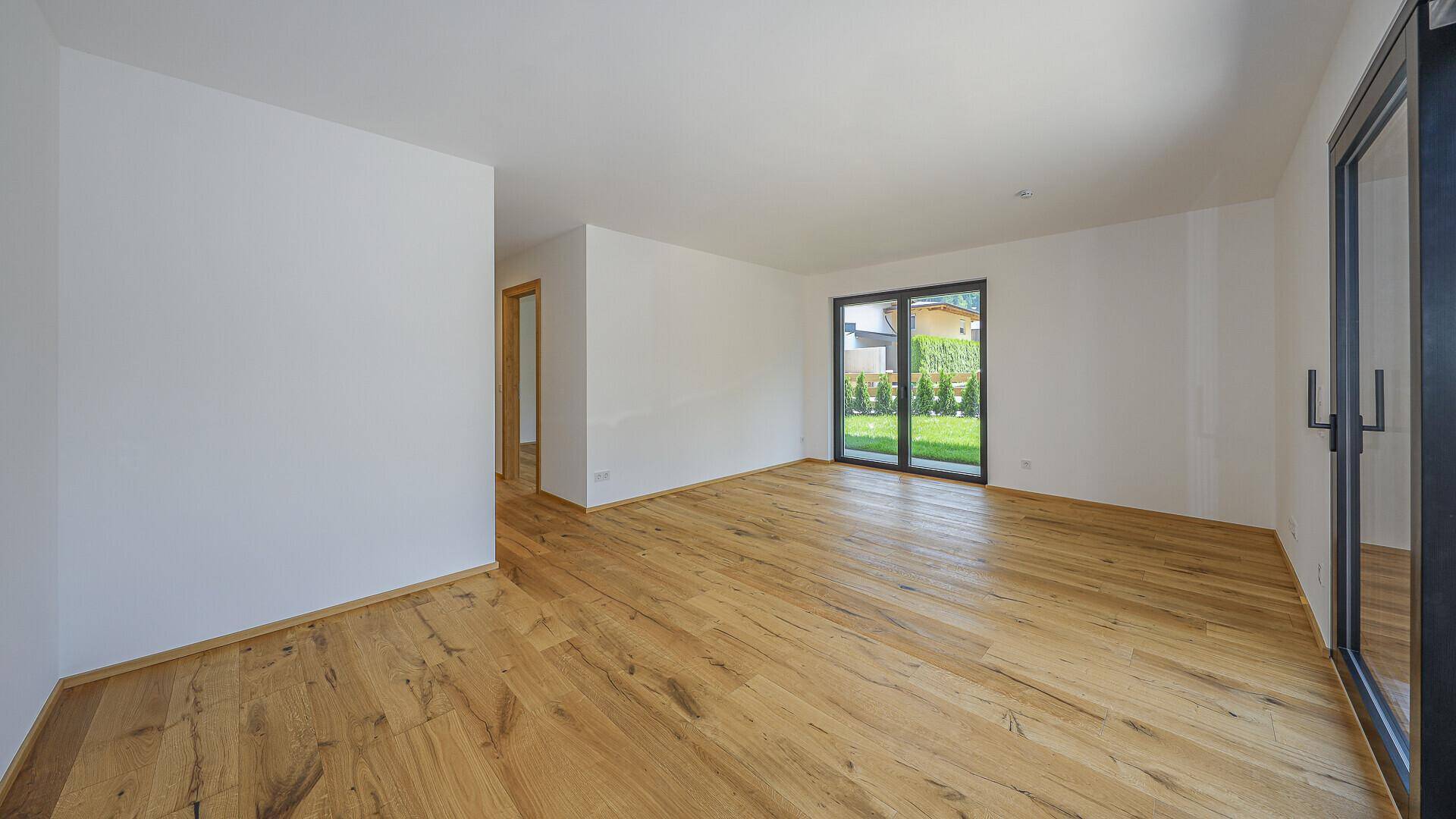 Leerer Wohnraum mit Holzboden und Blick ins Grüne durch die Terrassentür.