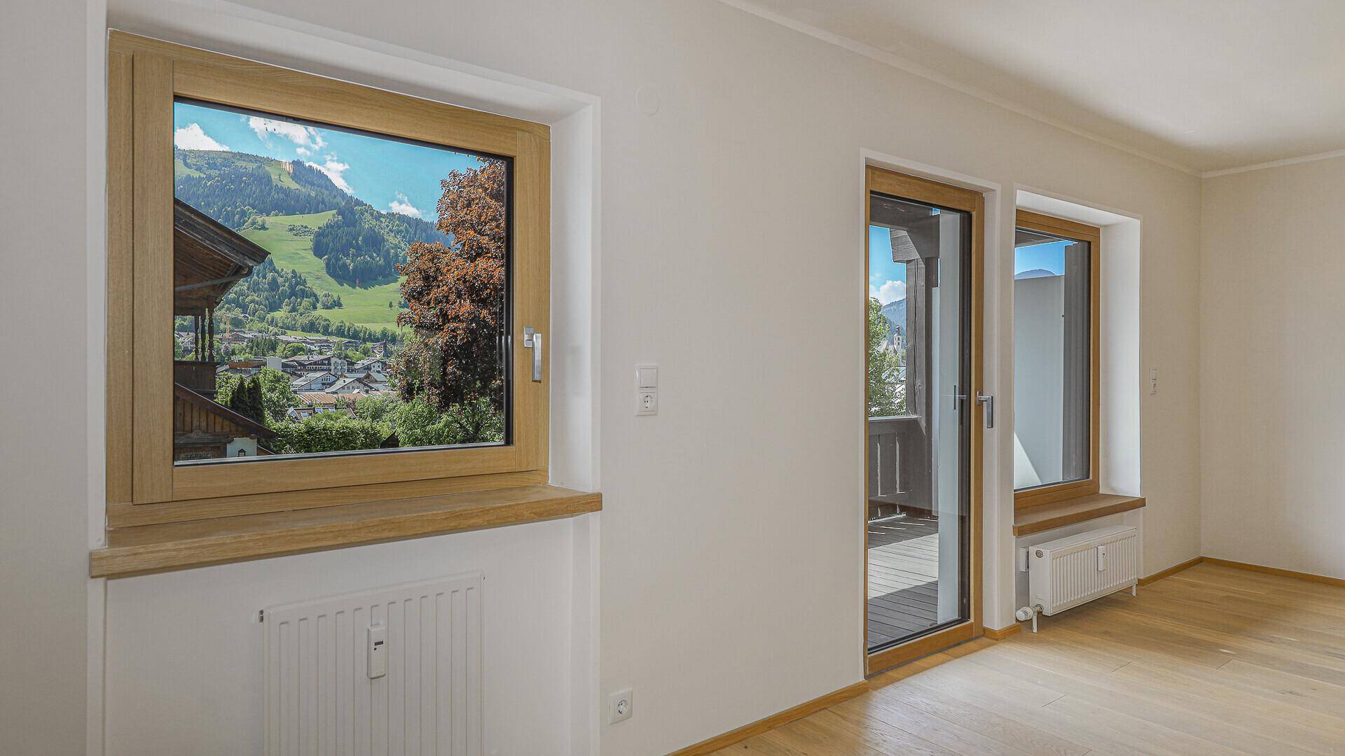 Heller Raum mit Bergblick durch das Fenster und Zugang zu einem Balkon mit Holzboden.