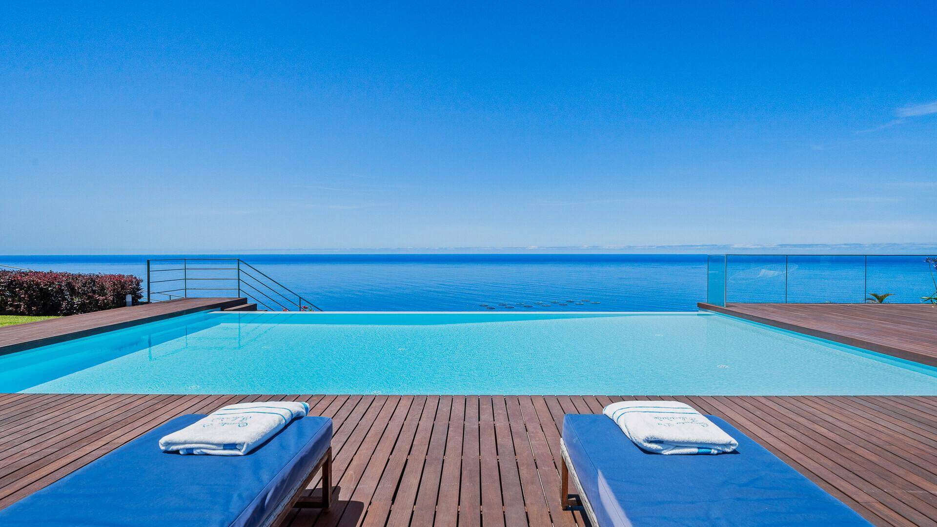 Entspannungsbereich am Infinity-Pool mit Holzdeck und Sonnenliegen, direkter Blick auf den Horizont des Meeres.