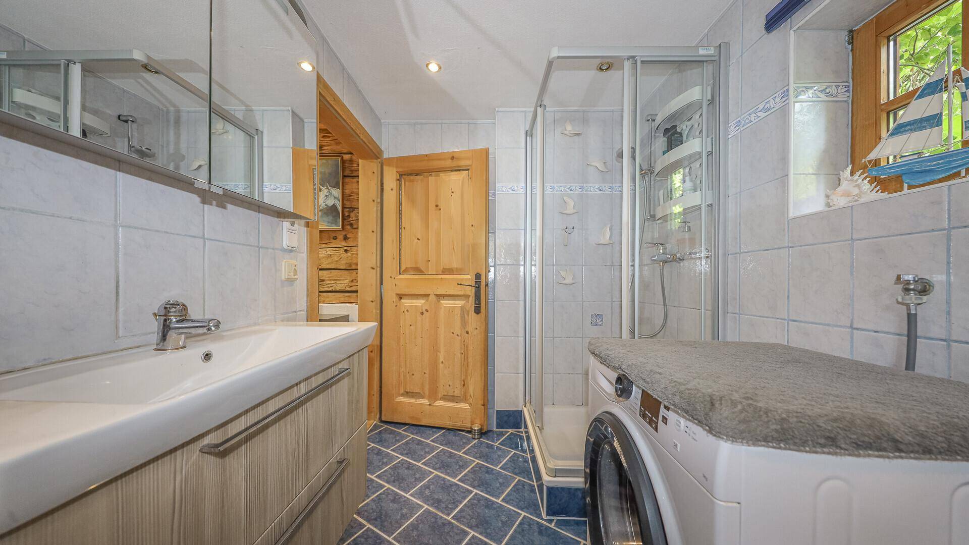 Badezimmer mit Dusche, Waschbecken und Waschmaschine, gefliest in hellen und blauen Tönen.