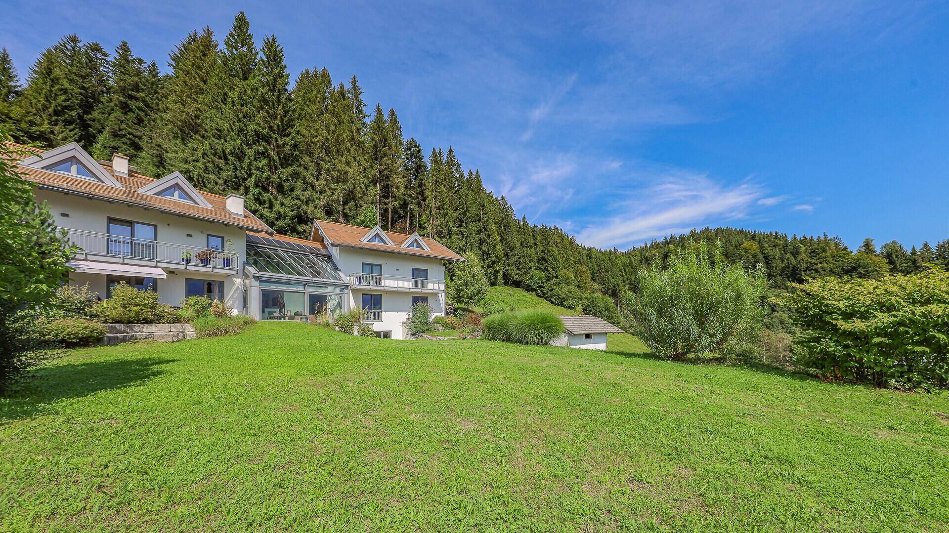 Ansicht des Anwesens von der weitläufigen Grünfläche mit Wald im Hintergrund und blauem Himmel.