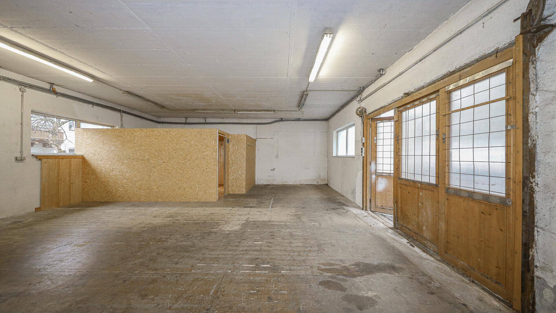 Großer Lagerraum mit Betonboden, mehreren Fenstern und provisorischen Holztrennwänden für verschiedene Bereiche.