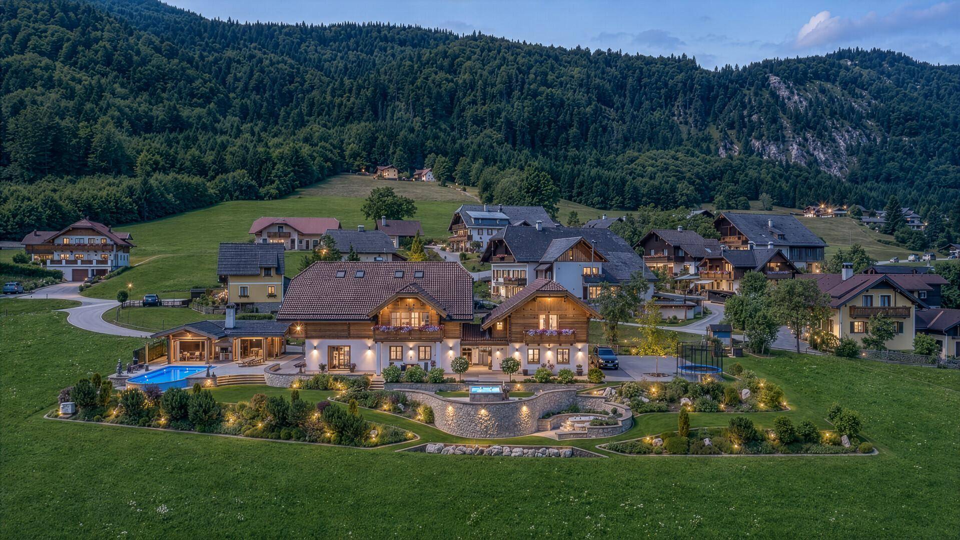 Abendliche Luftaufnahme eines beleuchteten Anwesens mit Pool, Garten und umliegender Natur.