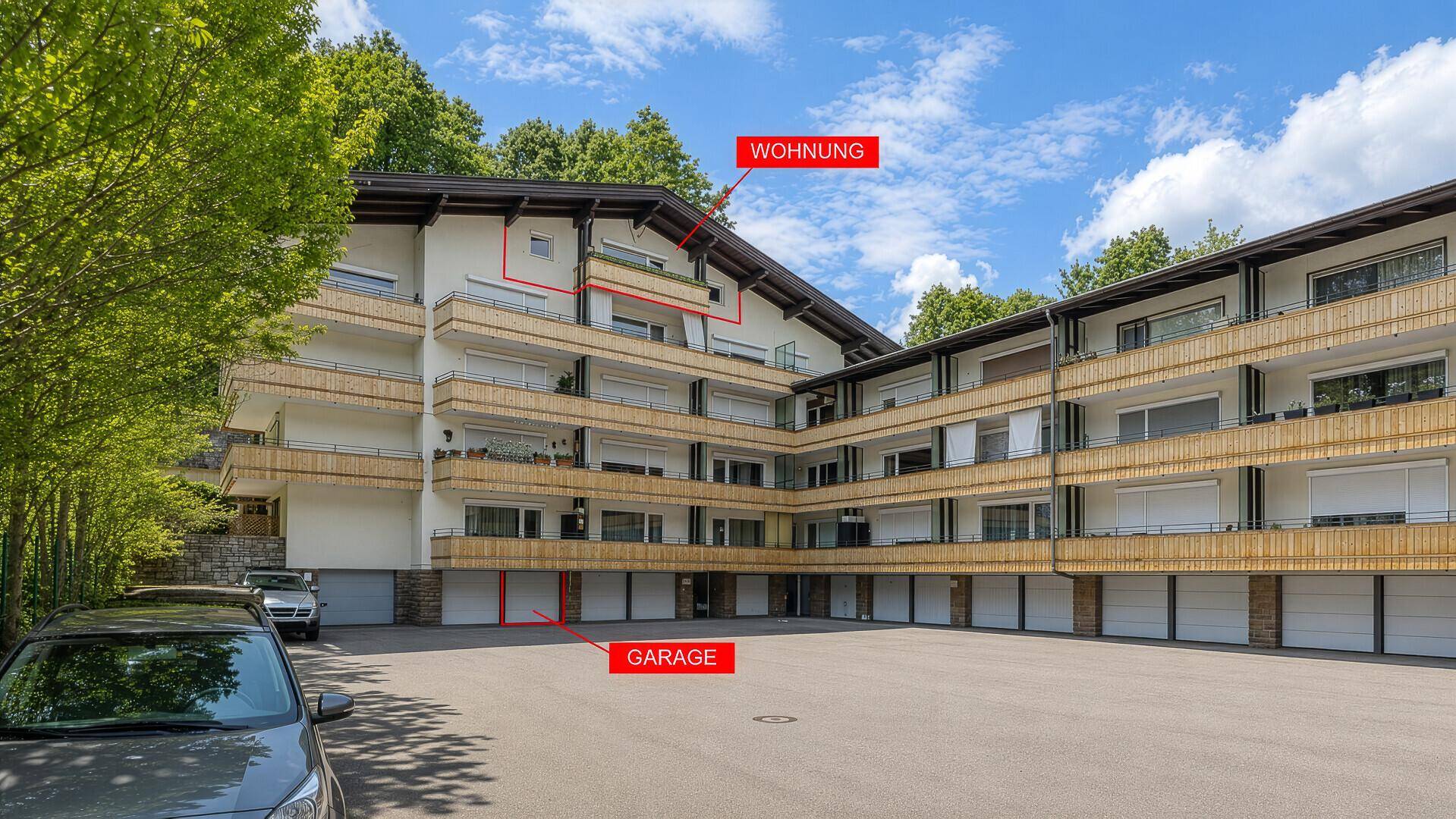 Mehrfamilienhaus mit Balkonen und Garagen, umgeben von Bäumen und Parkplätzen.